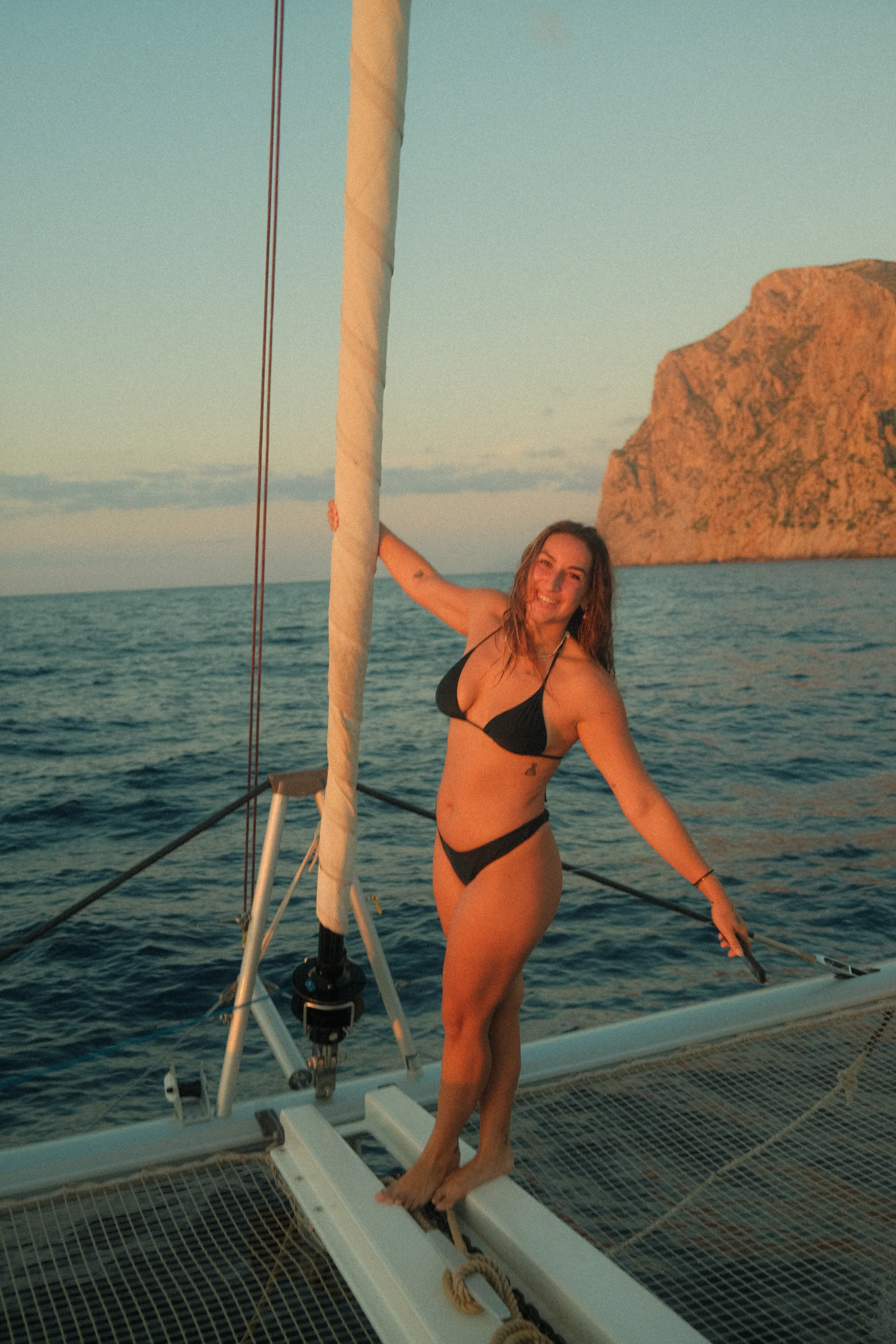 A woman in a black bikini standing on a catamaran with a rocky island in the background during sunset.