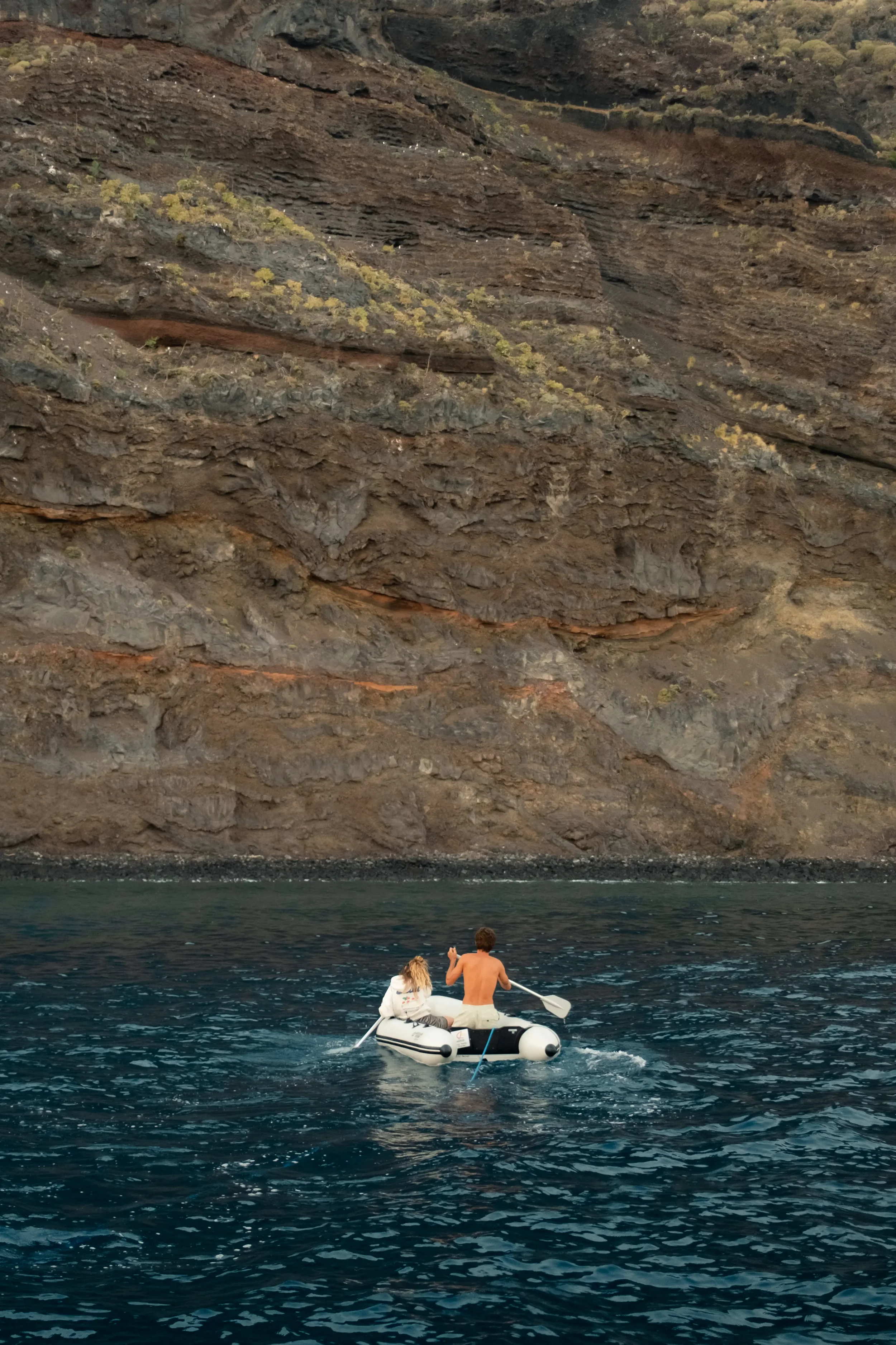 Two people are riding a paddle boat on dark blue water near a large rocky cliff.
