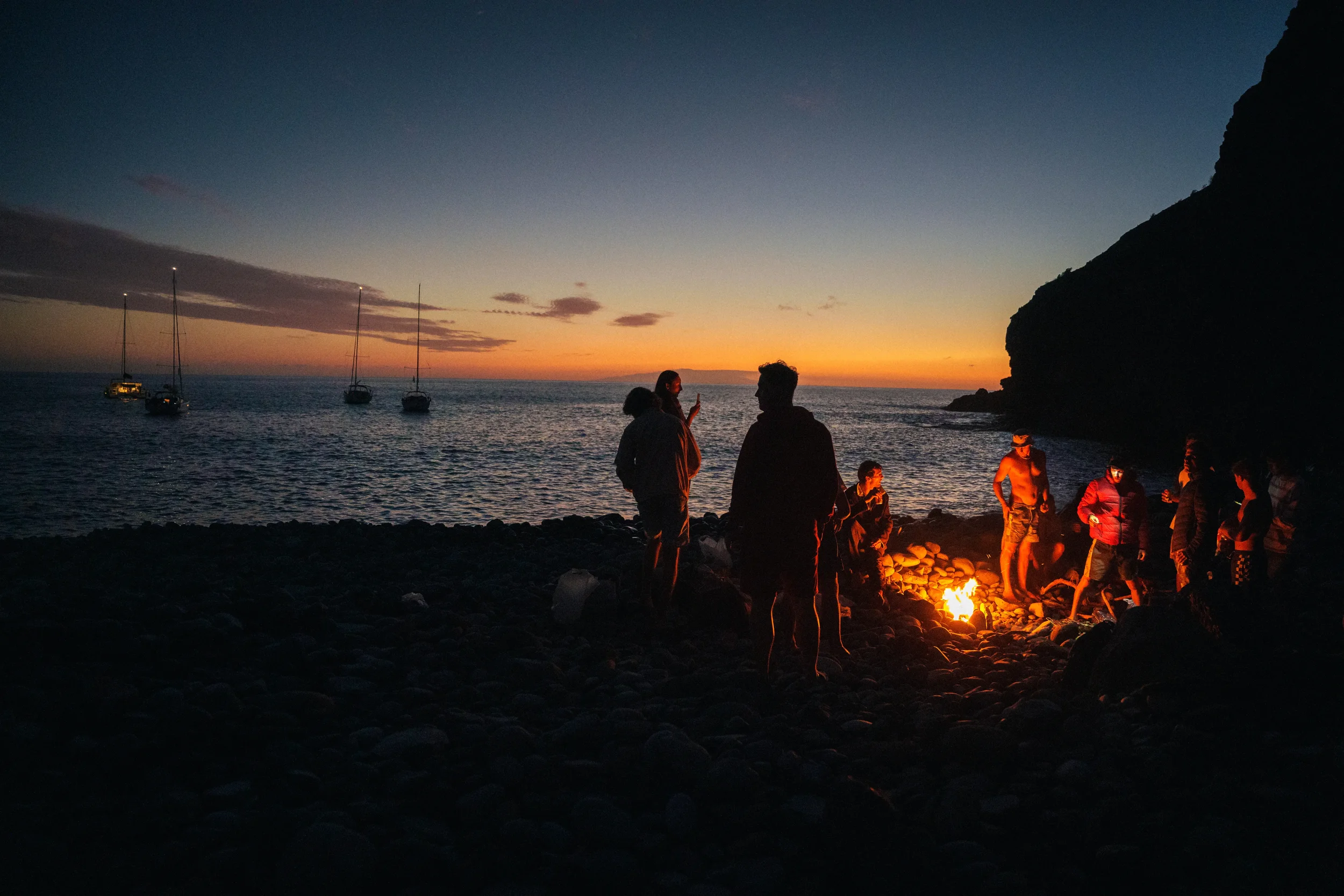 People gathered around a campfire on a rocky beach at sunset, with sailboats anchored in the water and a large rocky cliff on the right side.