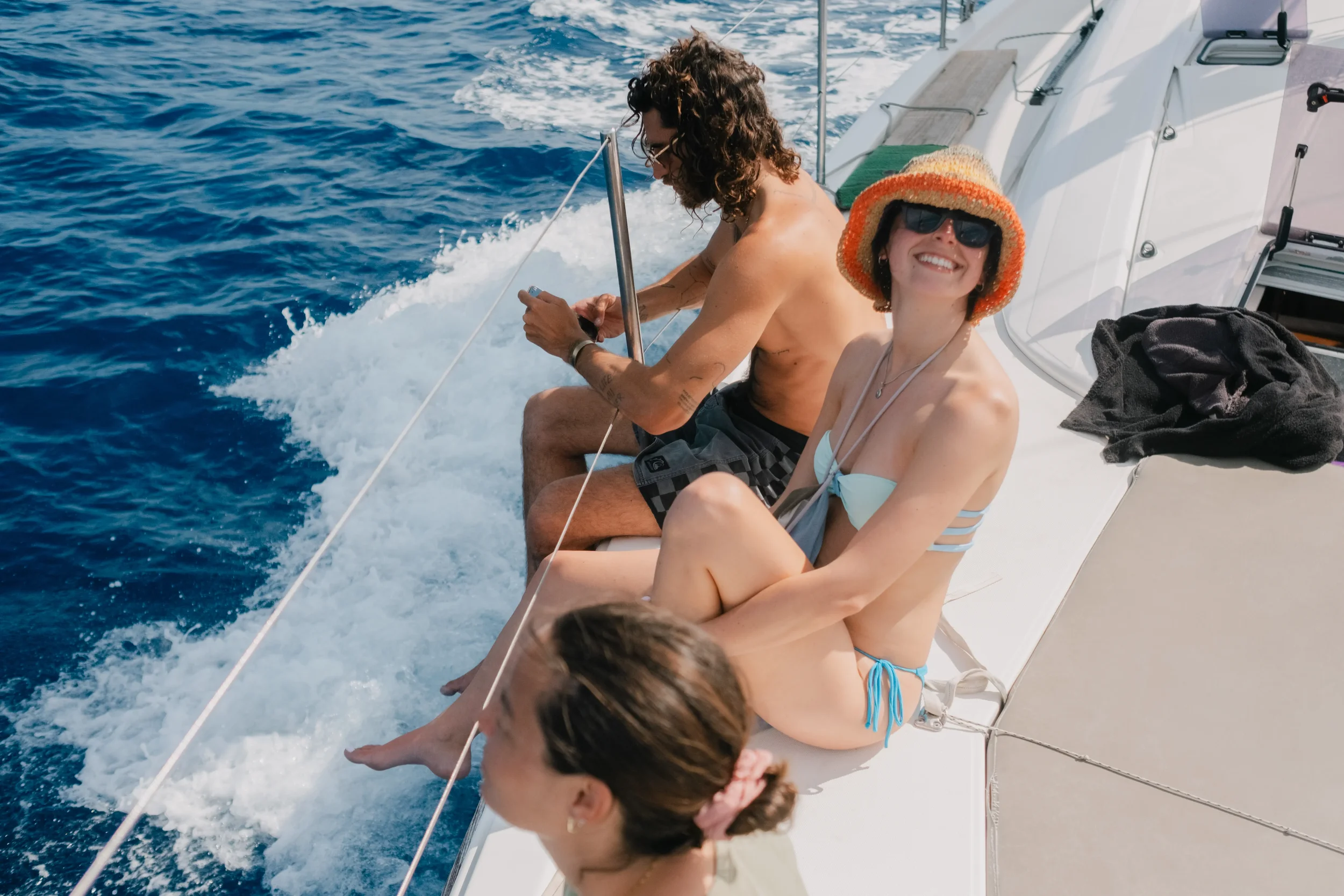 Two people sitting on the edge of a boat, enjoying the ocean, with one smiling and wearing sunglasses and a straw hat, the other looking down, both in swimwear.