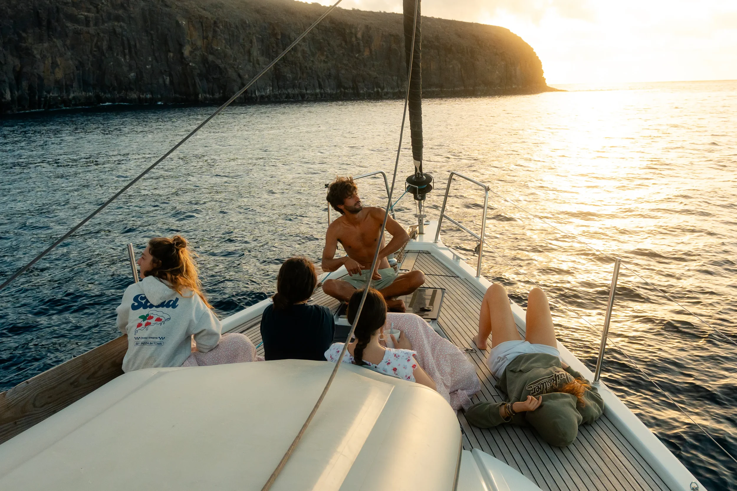 A group of people relaxing on a boat during sunset, with a rocky cliff in the background and calm water surrounding them.