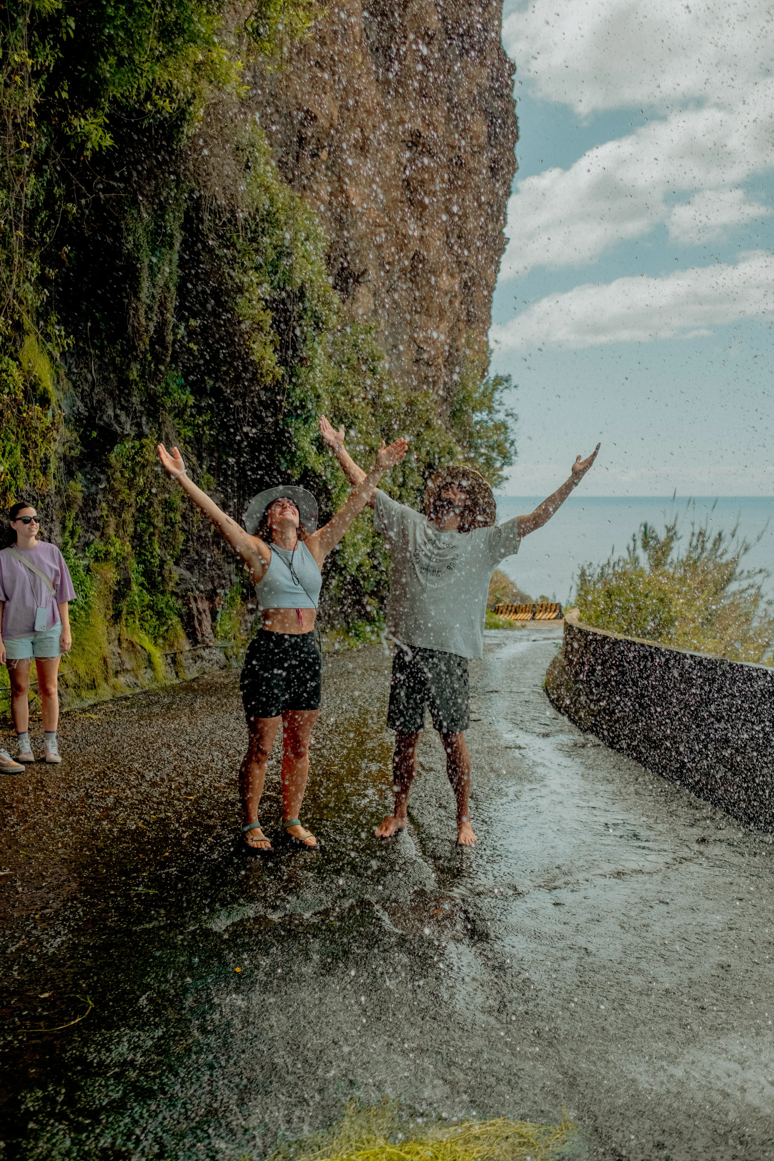 Three people standing on a rocky shore next to water, with two of them throwing water into the air and smiling, while a woman on the left looks on. There are trees and a rocky cliff in the background under a partly cloudy sky.