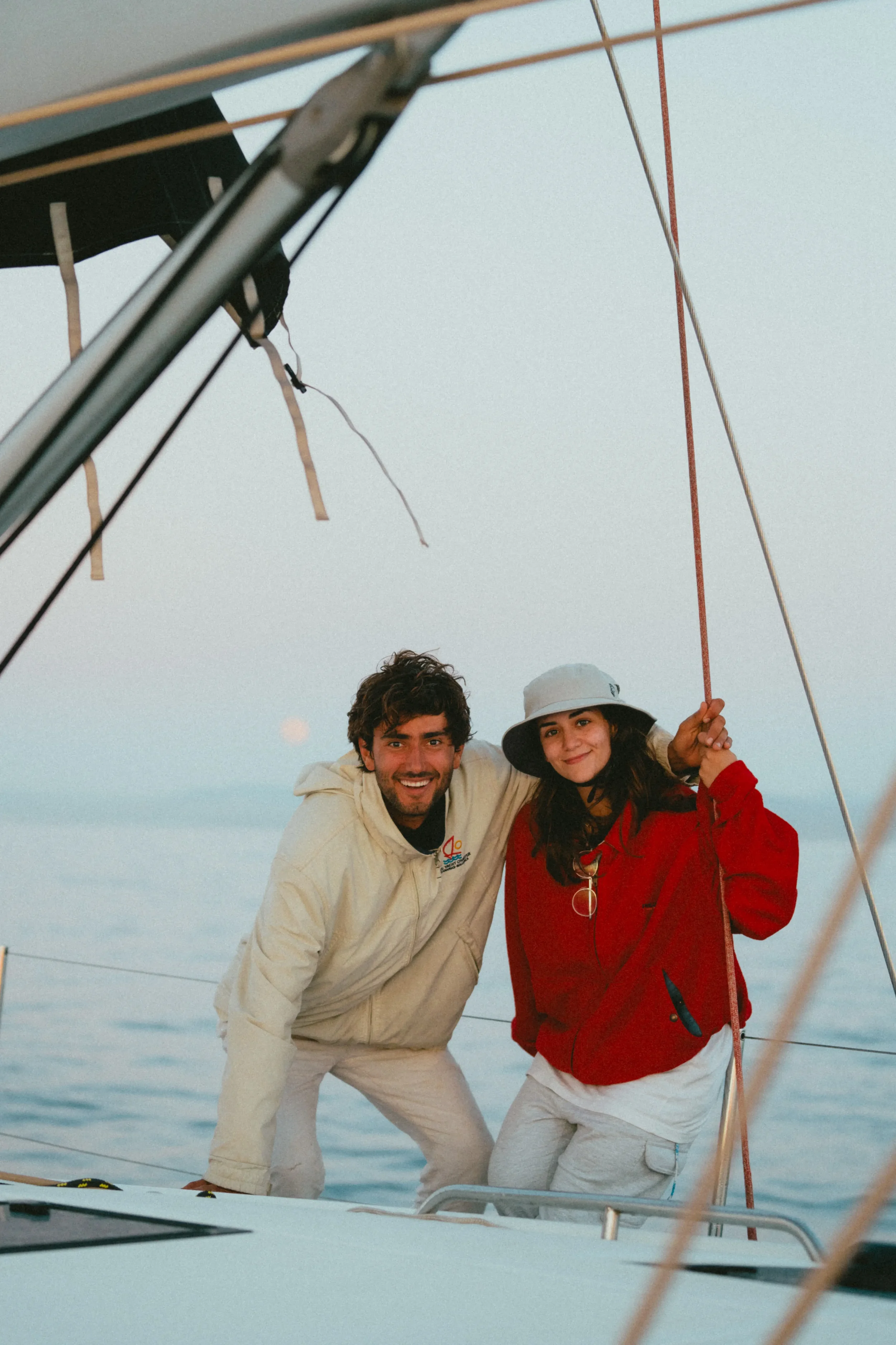 Two people, a man and a woman, aboard a boat, smiling and holding onto a rope, with a calm ocean and an overcast sky in the background.