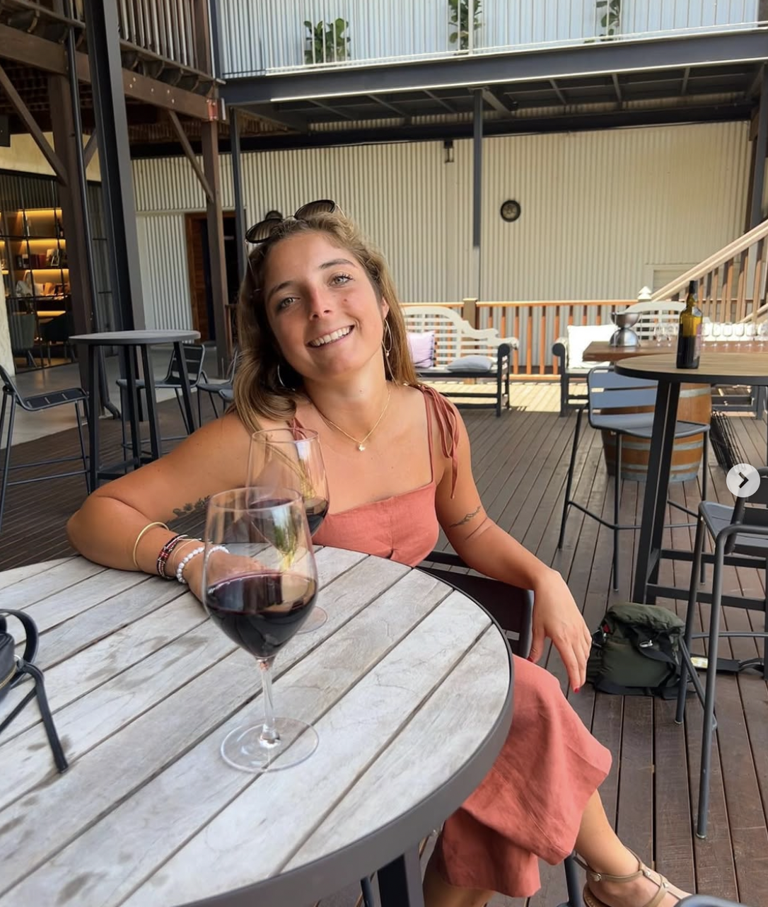 A woman sitting at a table outdoors, smiling with two glasses of red wine, in a modern patio setting.