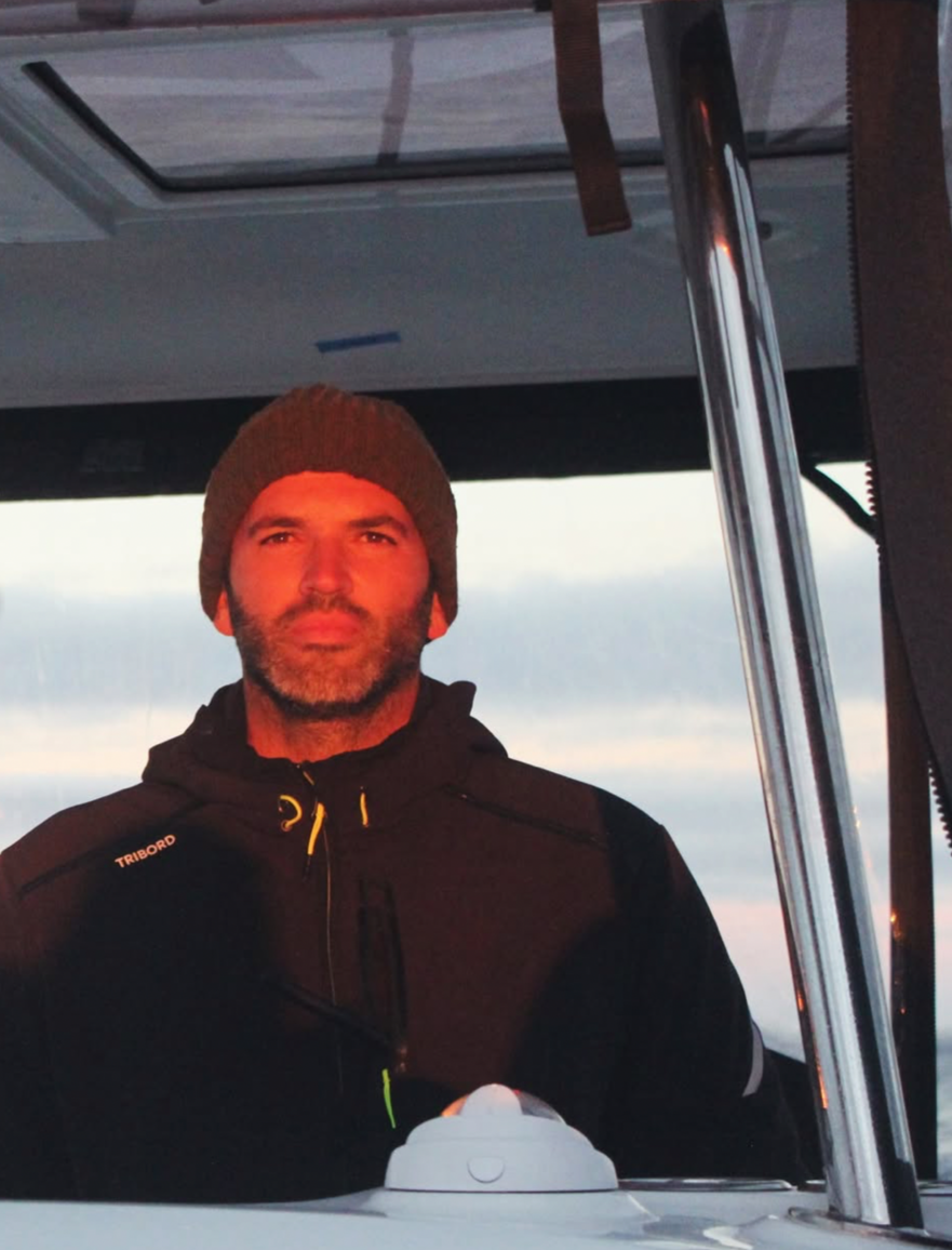 A man with a beard and mustache wearing a gray knit beanie and black jacket, sitting at the helm of a boat during sunset with a cloudy sky in the background.