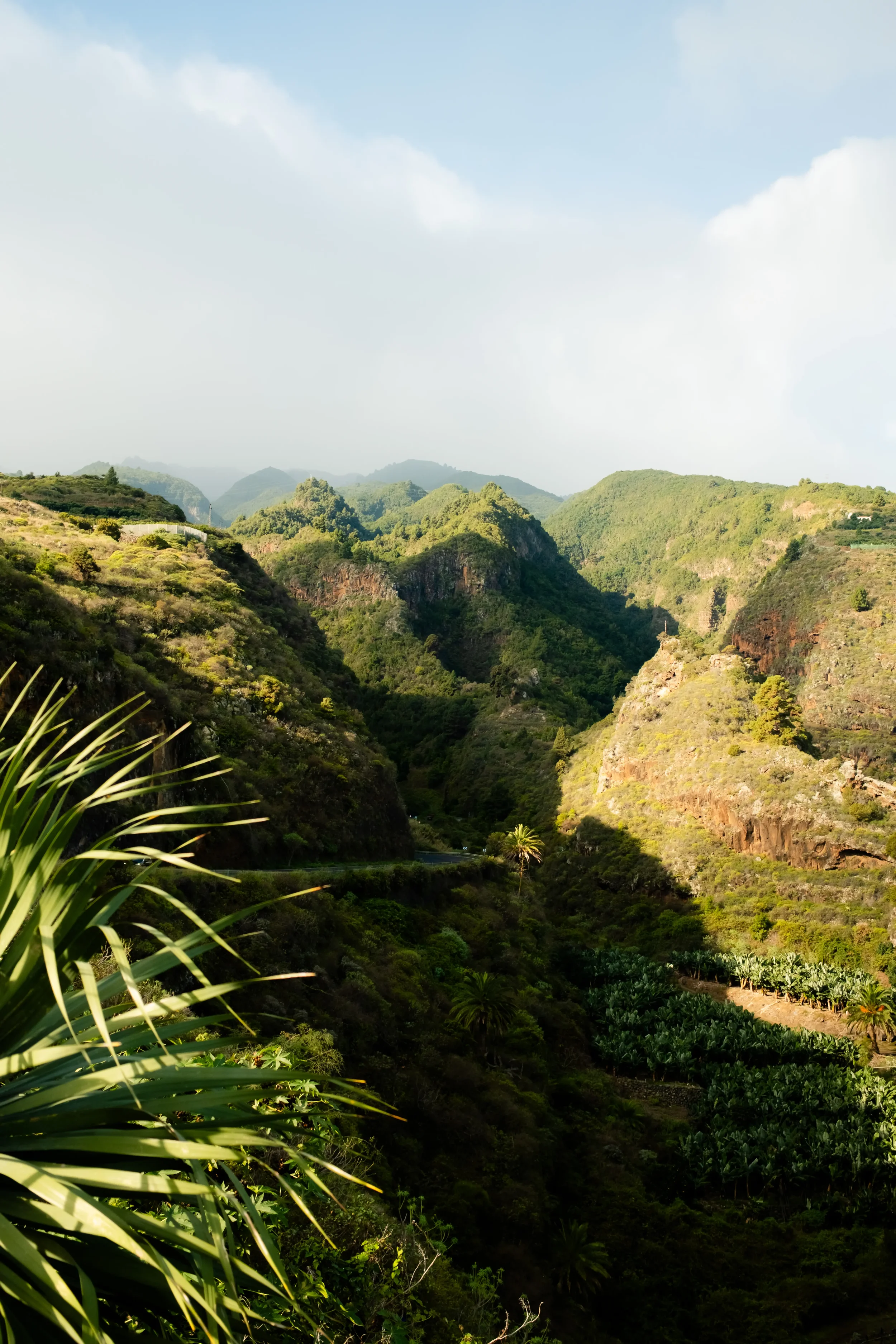 Lush green mountains and valleys with a winding road in the foreground, under a partly cloudy sky.