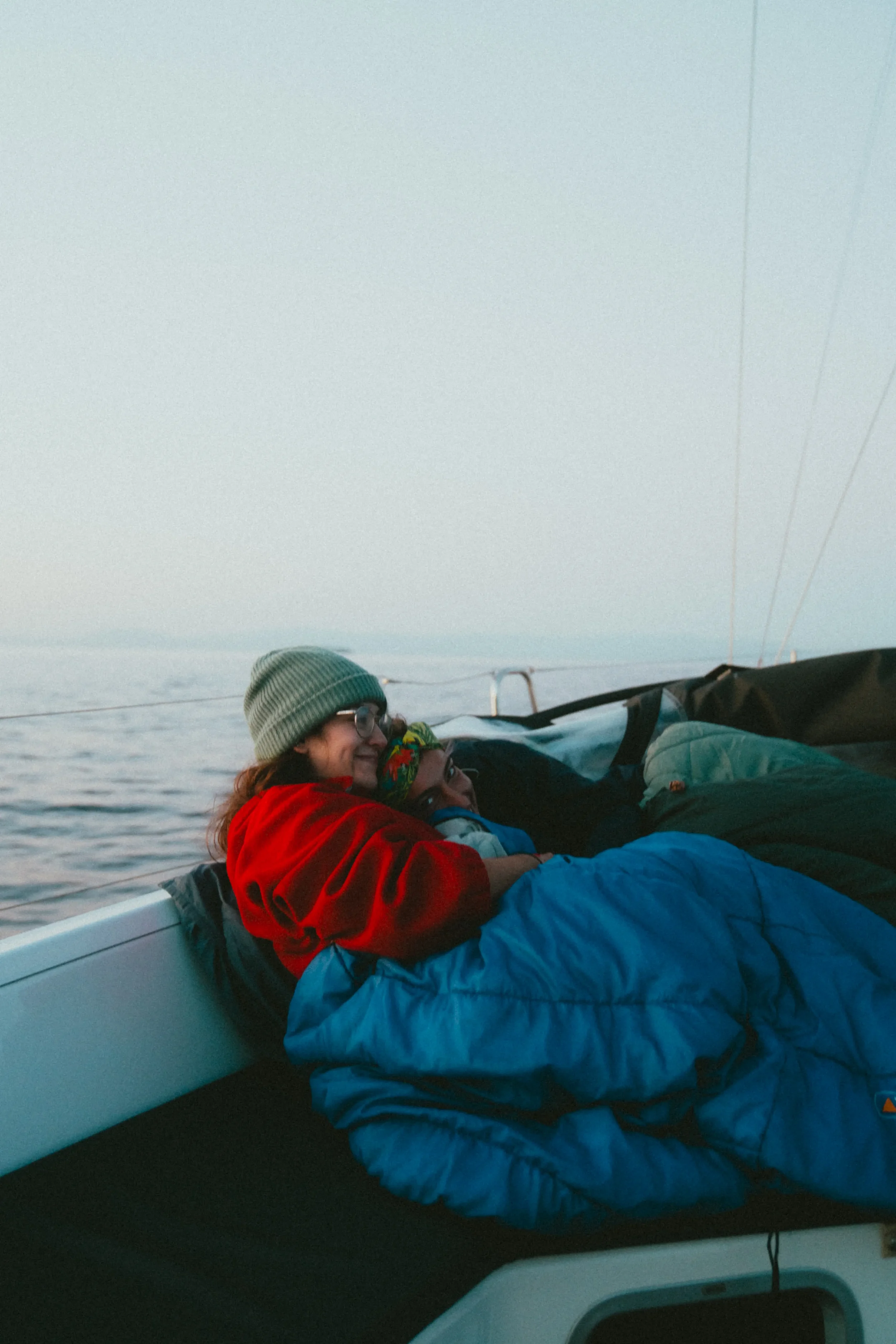 Two women in warm clothing, one wearing a green beanie and glasses, smiling while lying close together on a boat during sunset or sunrise with water and sky in the background.