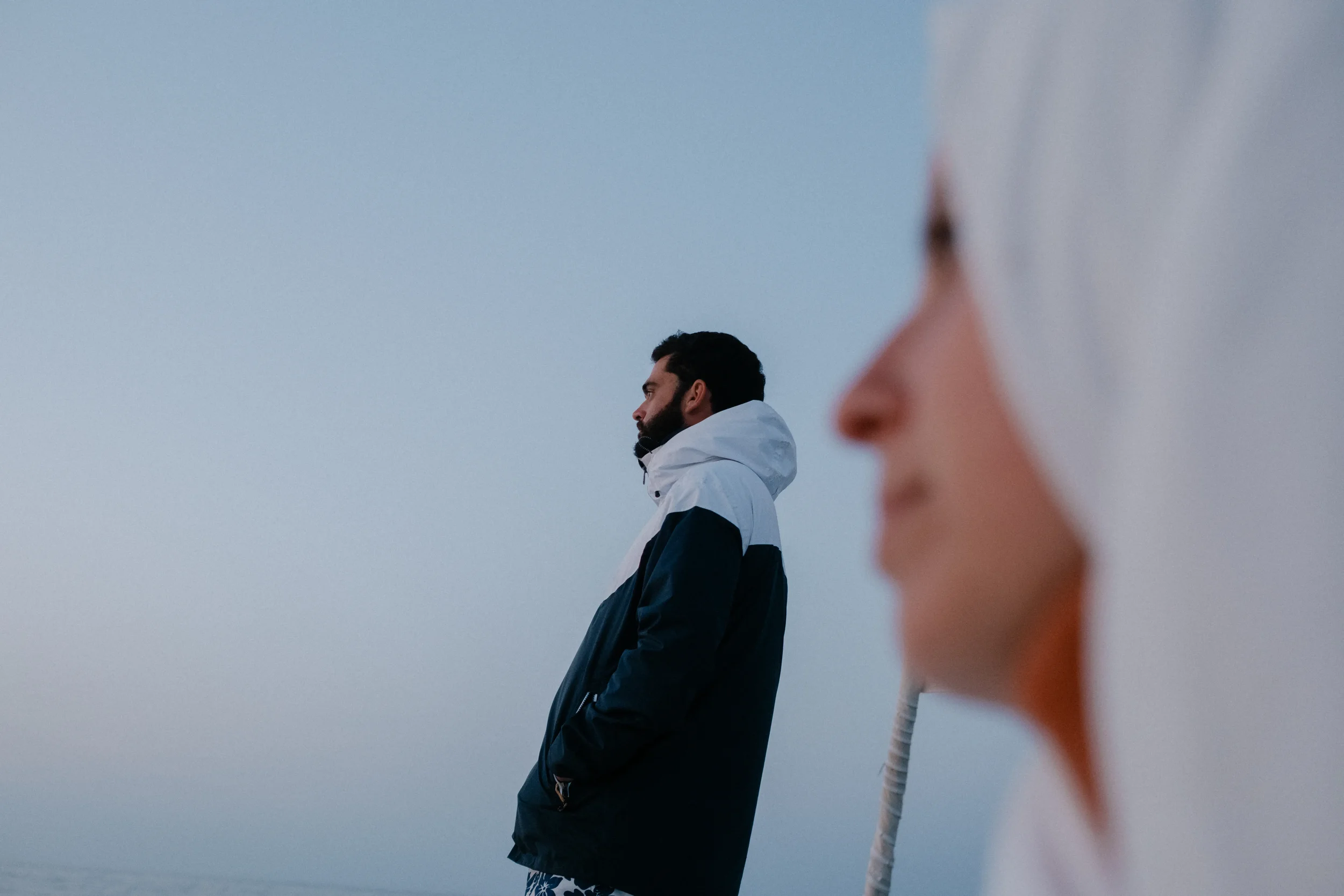 A man with dark hair and a beard wearing a black jacket, standing with his hands in his pockets, looking to the left, against a pale blue sky. Part of a person's face is visible in the foreground on the right side of the image, slightly blurred.