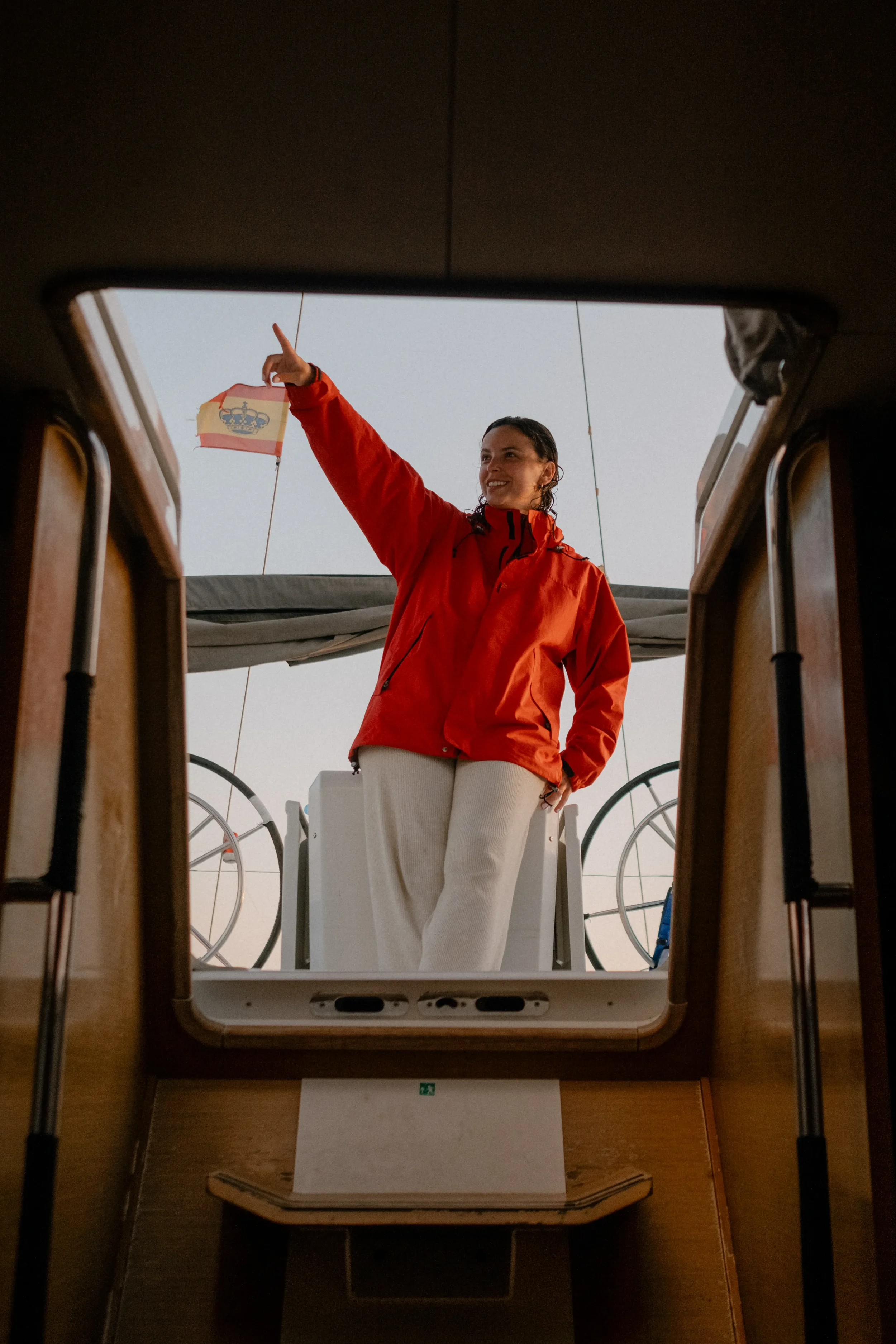 Smiling woman in a red jacket standing on a boat, pointing upward, seen through a hatchway with a flag featuring a crown and a red and white background in the background.