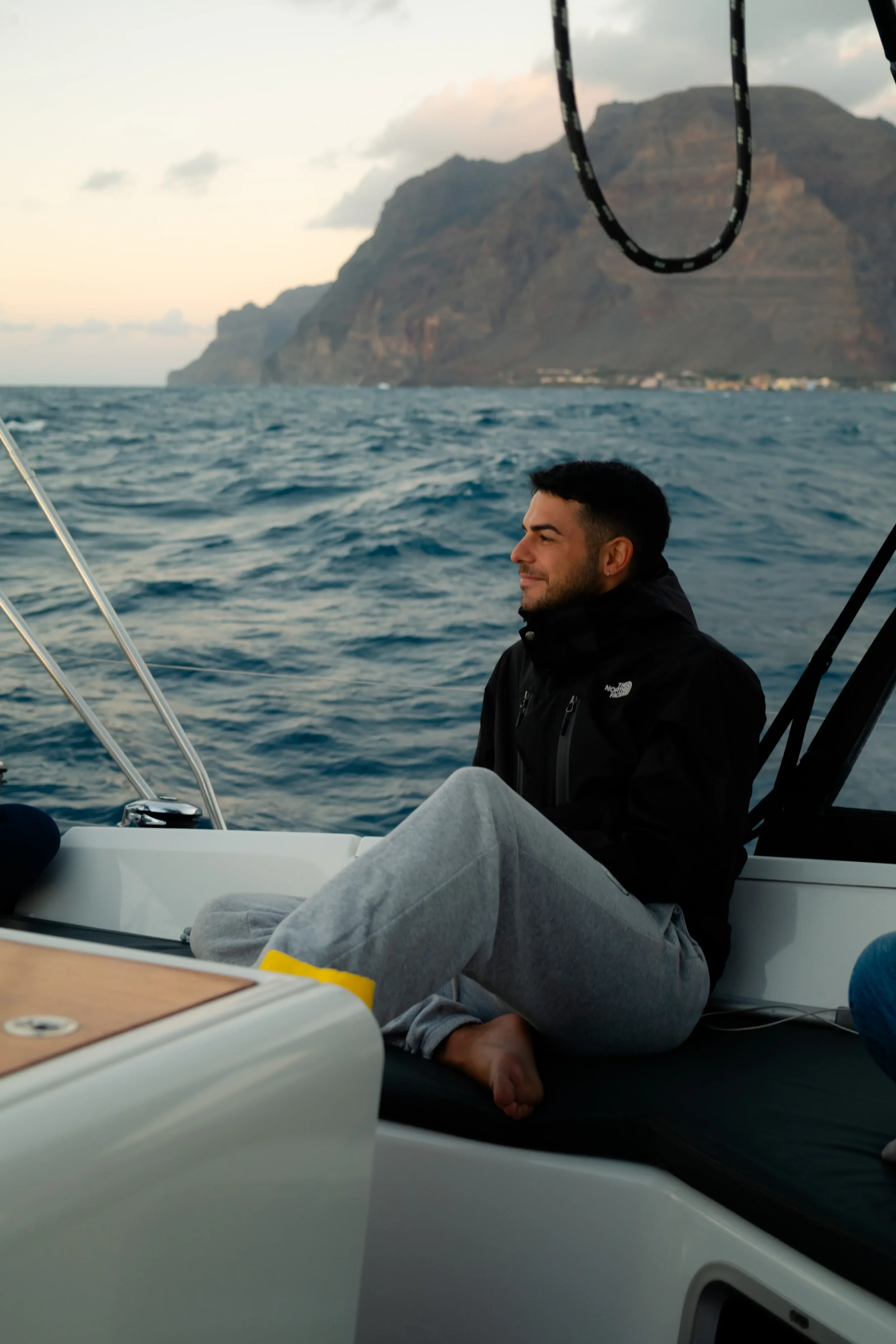 A man sitting on a boat, smiling, with mountains and water in the background during what appears to be sunrise or sunset.