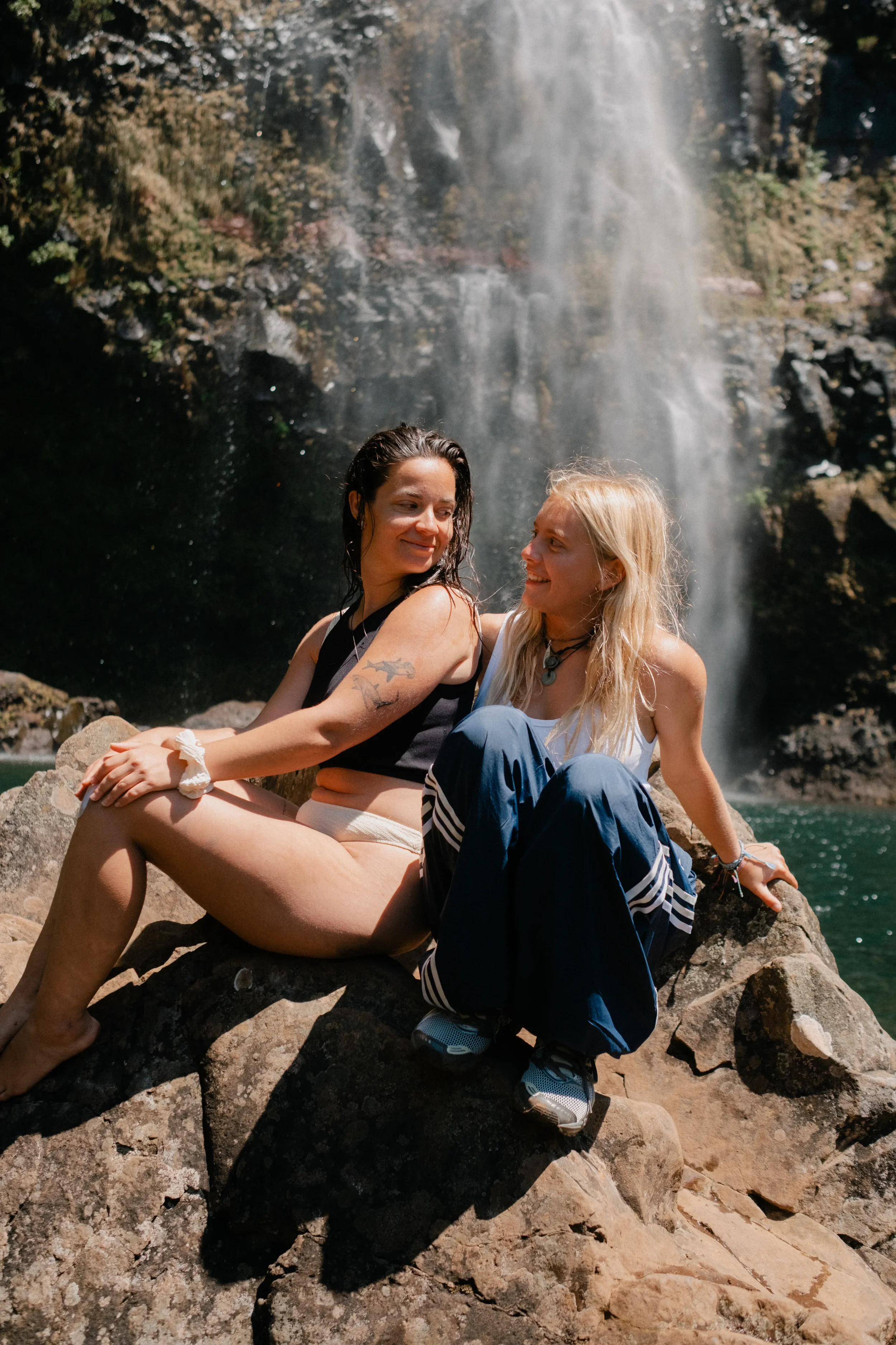 Two women sitting on rocks in front of a waterfall, smiling and looking at each other, with water spray and greenery in the background.