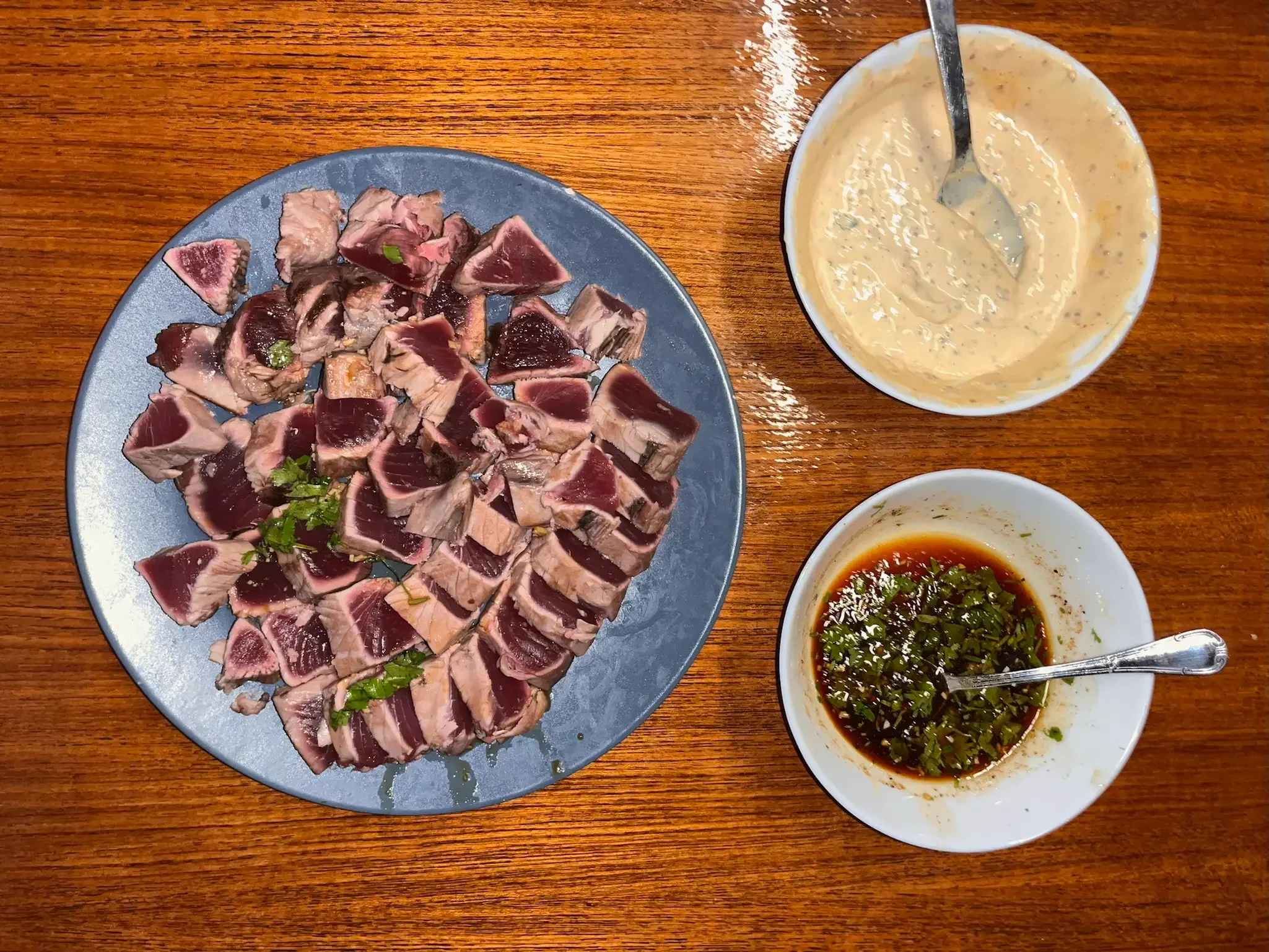 Sliced raw tuna served on a blue plate with some green herbs, accompanied by three small bowls of sauce and condiments on a wooden table.