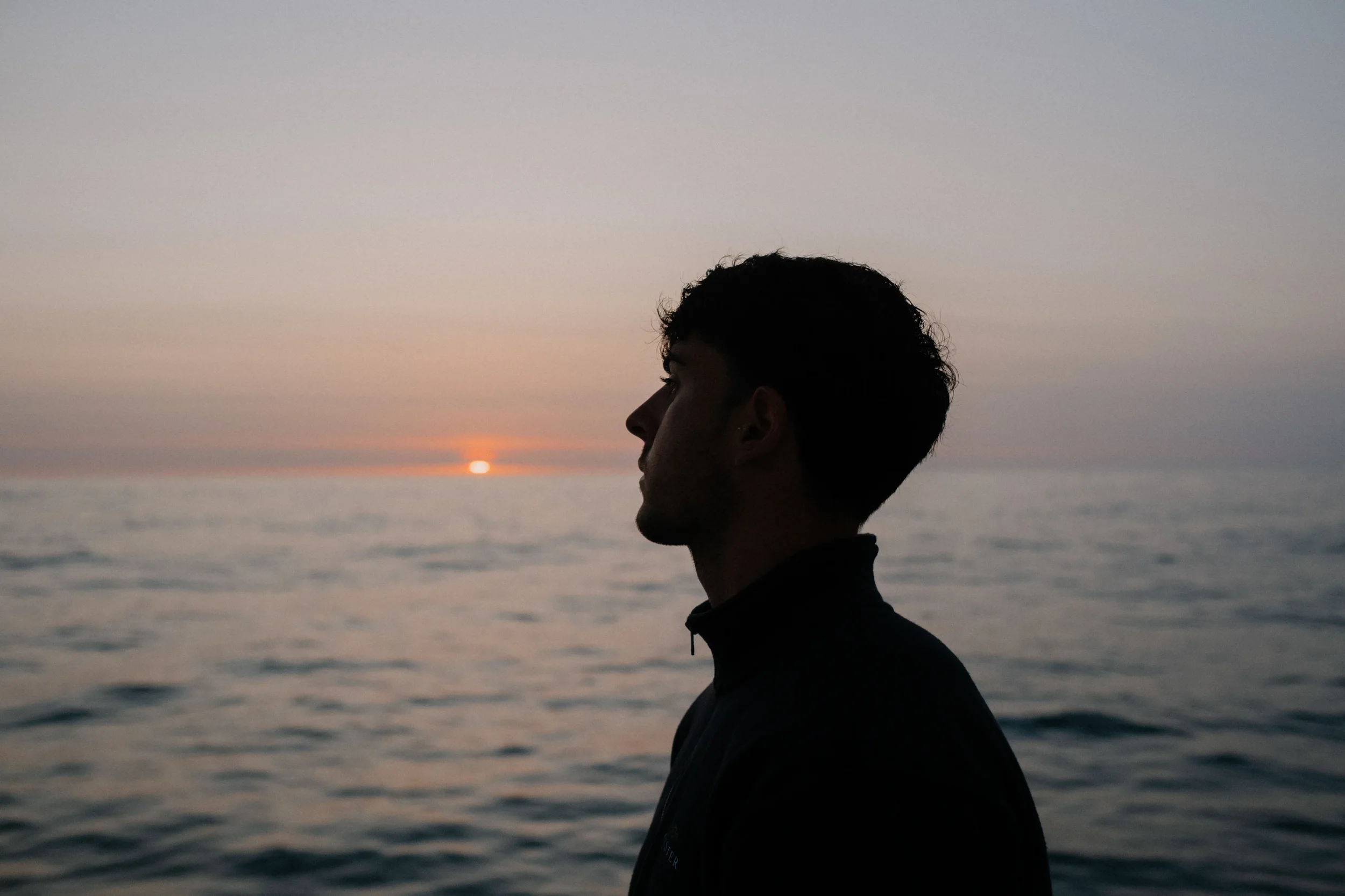 Silhouette of a person with short curly hair looking to the left, standing by the ocean during sunset with a colorful sky.