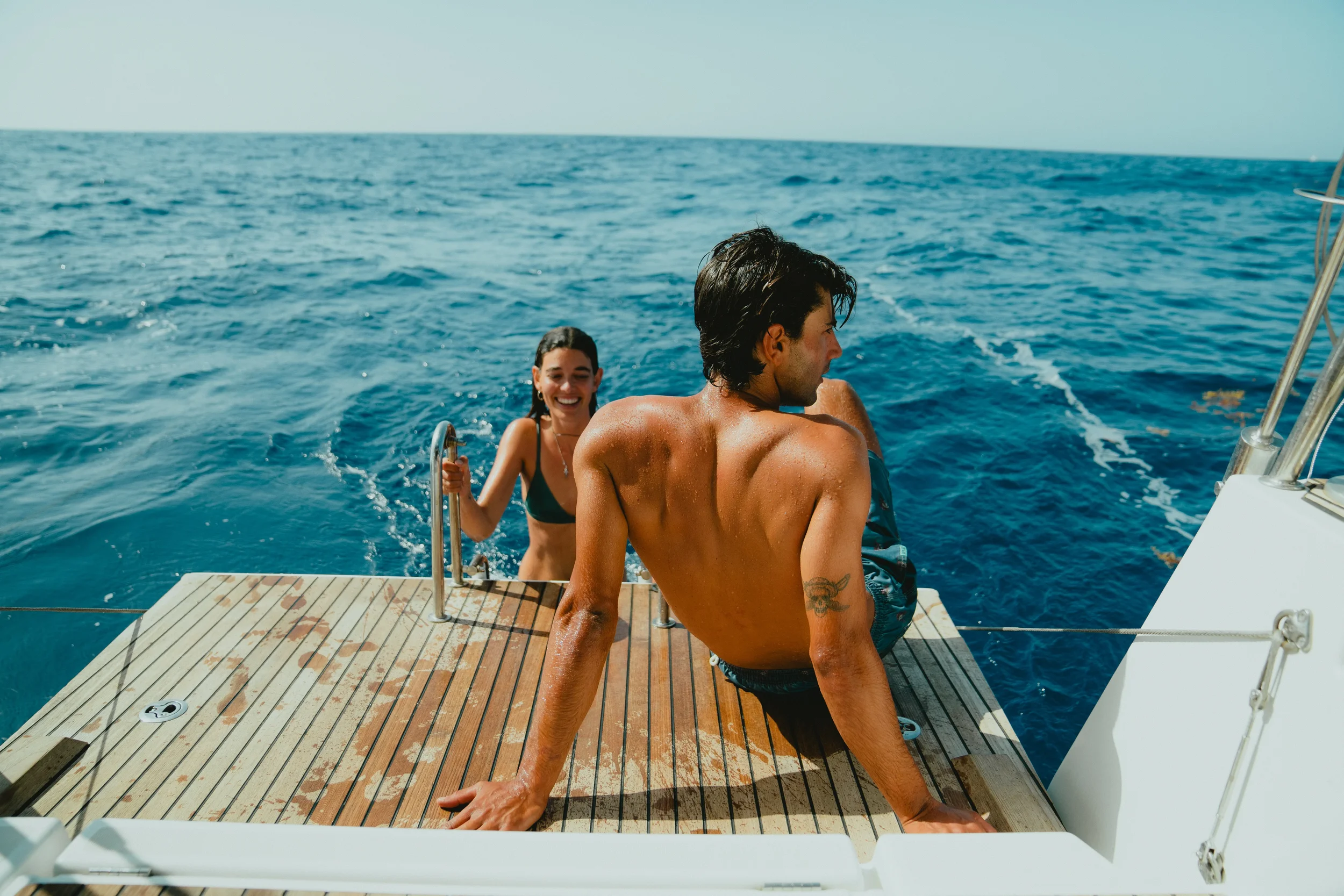 Two smiling people on a boat, with a man shirtless and a woman in a dark green swimsuit, in the open ocean under a clear sky.