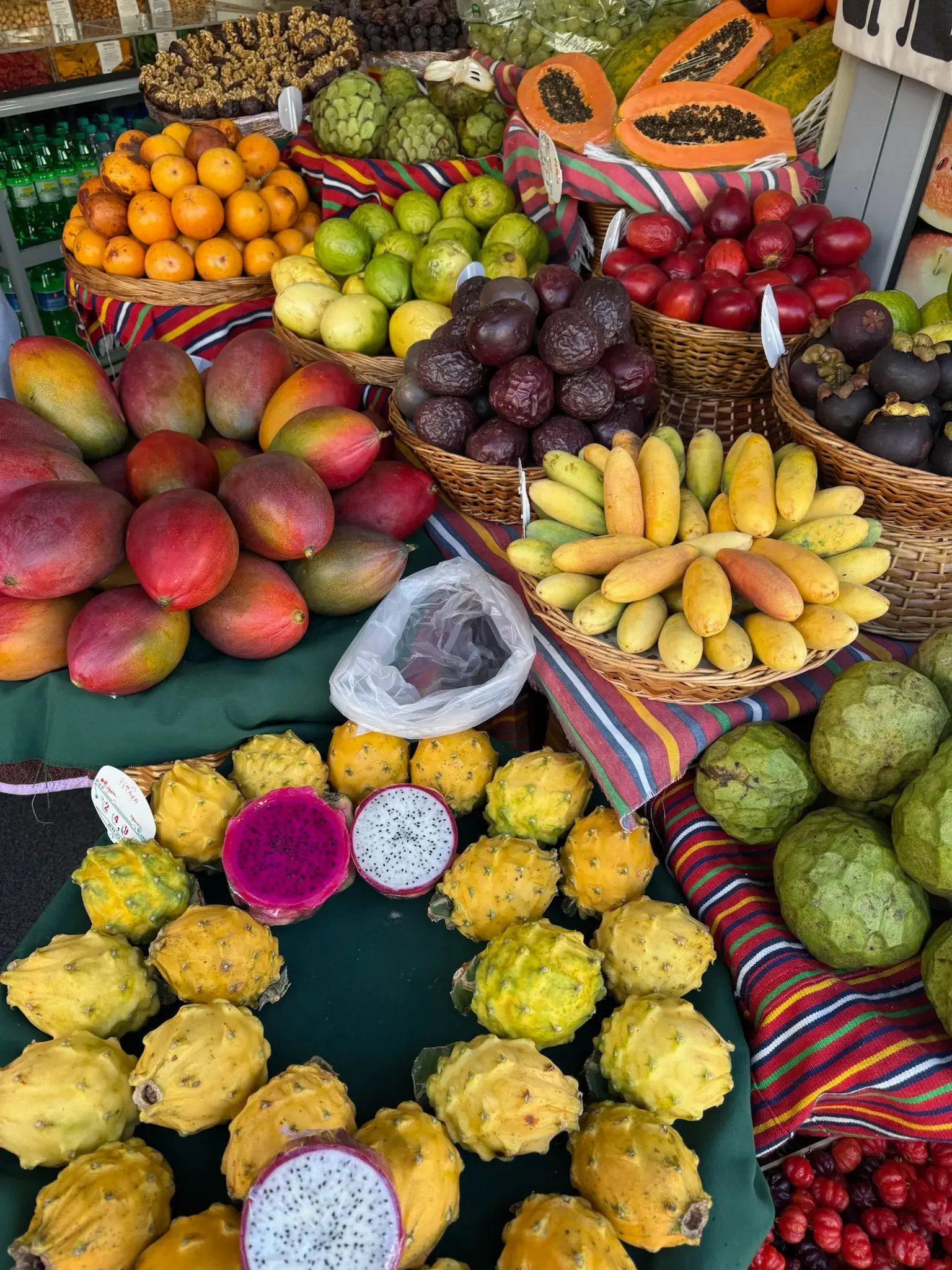 Display of various tropical fruits at a market stand, including dragon fruits, papayas, passion fruits, apples, mangoes, and other assorted fruits in baskets and on a striped tablecloth.
