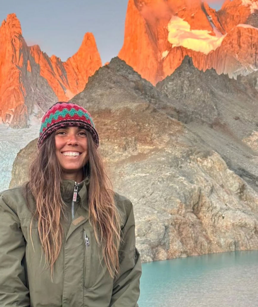 A woman smiling in front of a mountain landscape with orange and gray peaks, a lake at the base, and snowy patches.
