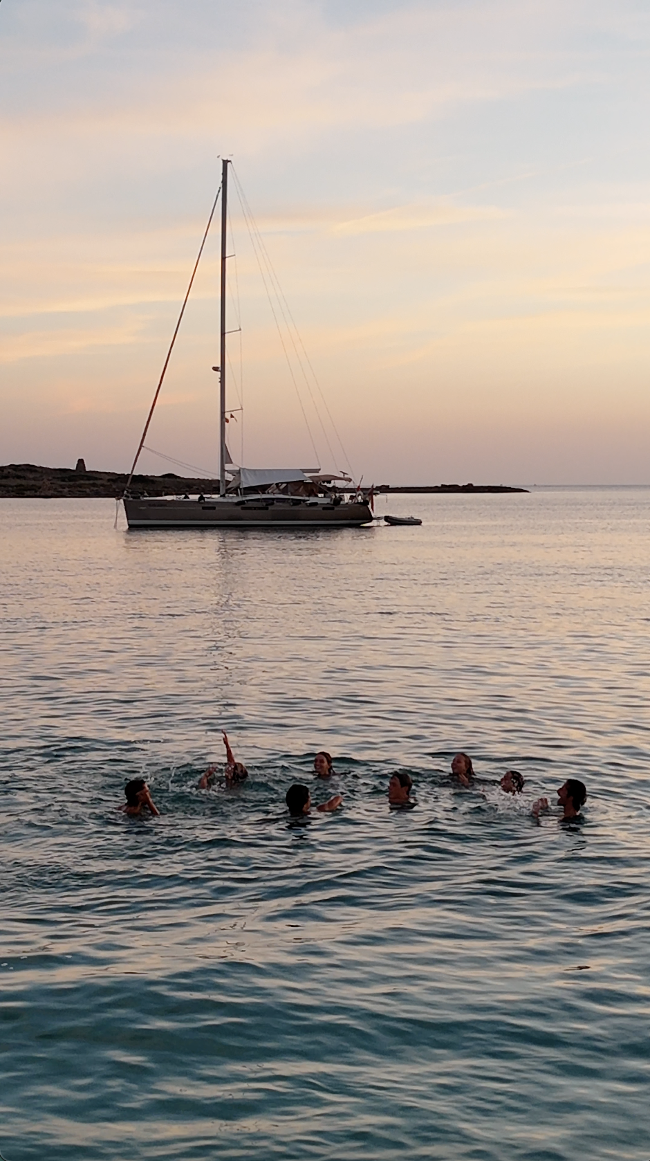 Group of people swimming in the water near a sailboat during sunset.