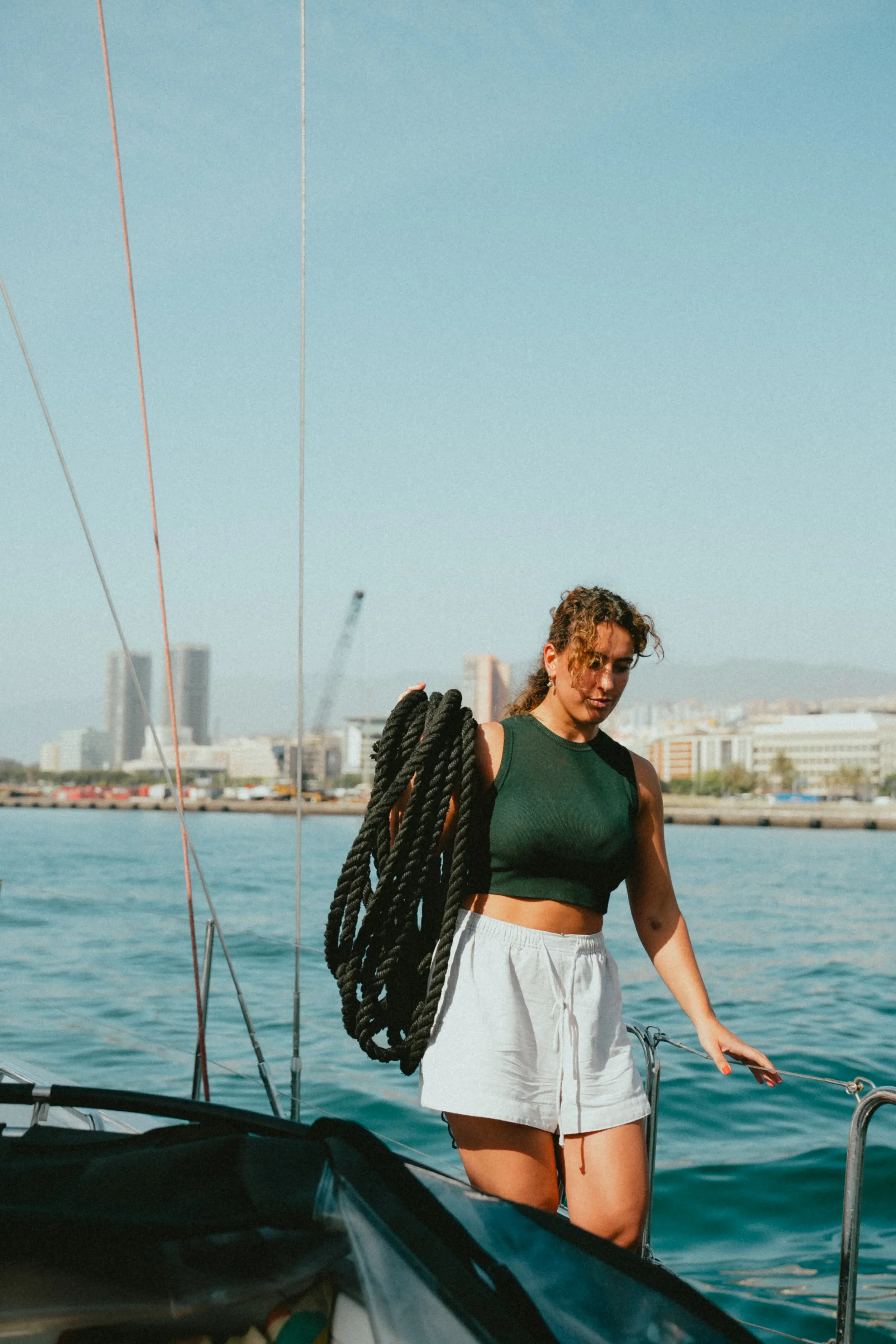 A woman with curly hair wearing a green sleeveless top and white shorts on a boat, holding onto the boat railing with a city skyline in the background.