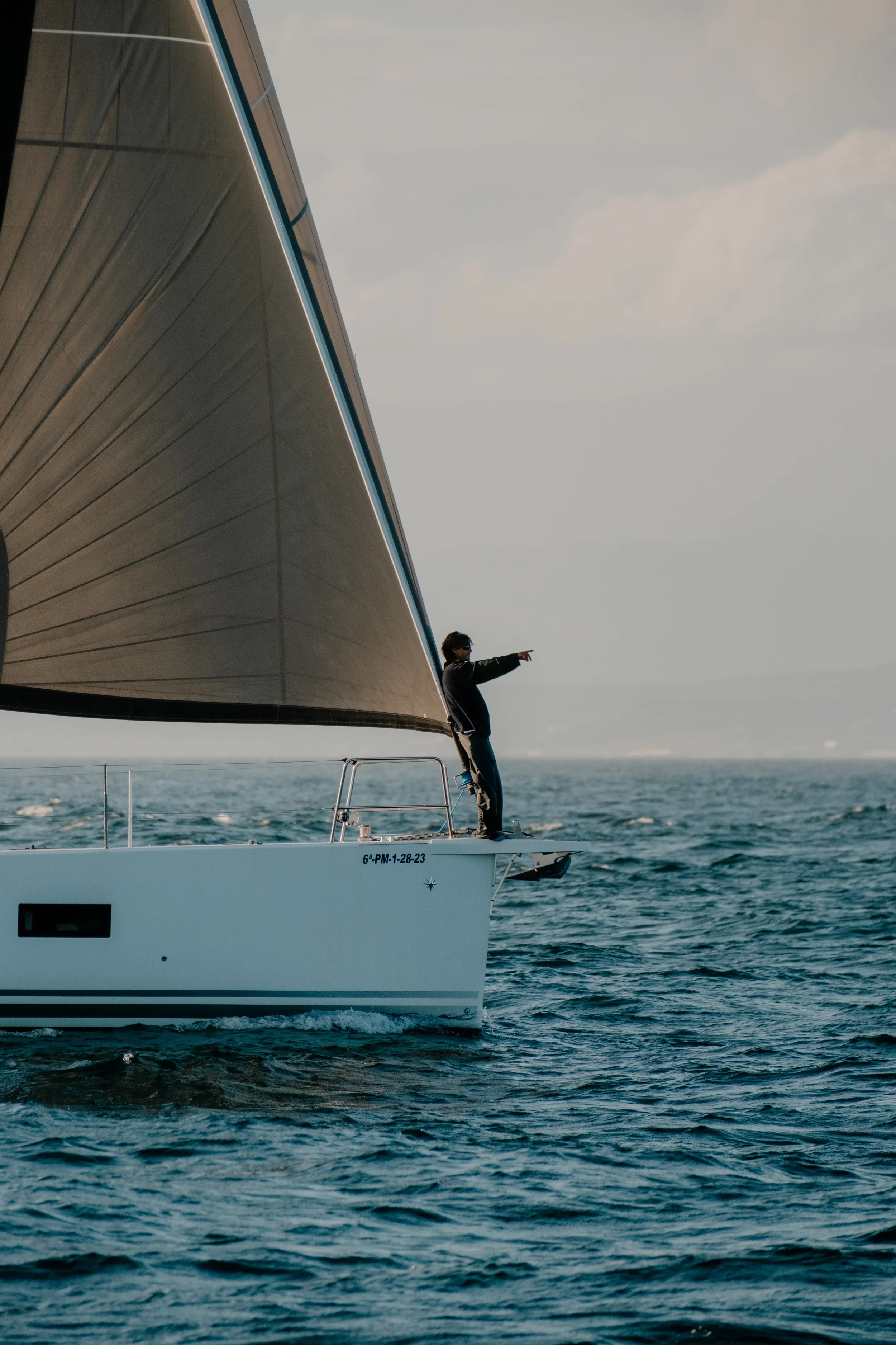 A person standing on the bow of a sailboat with its arms stretched out, sailing on open water during sunset.
