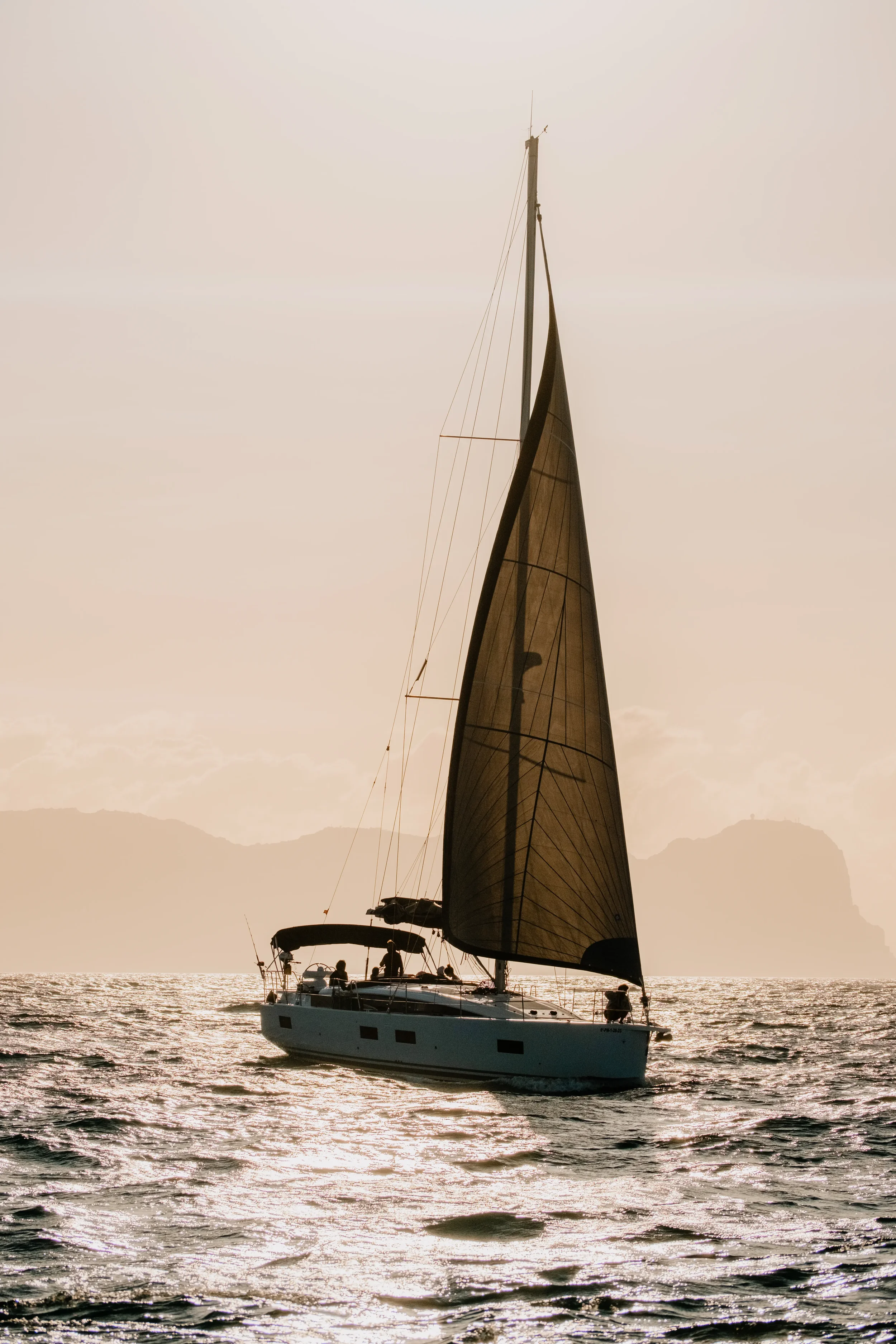 A sailboat with black sails and a white hull sailing on water during sunset, with a few people on board, and mountains in the background.