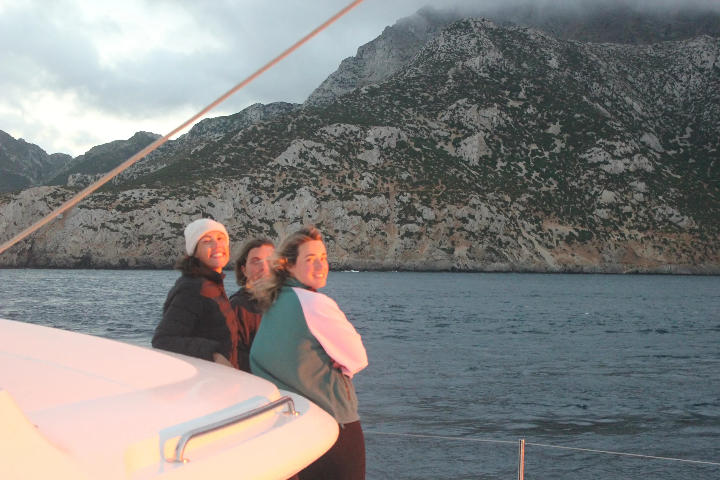Three people on a boat smiling at the camera, with a mountainous coastline and cloudy sky in the background.