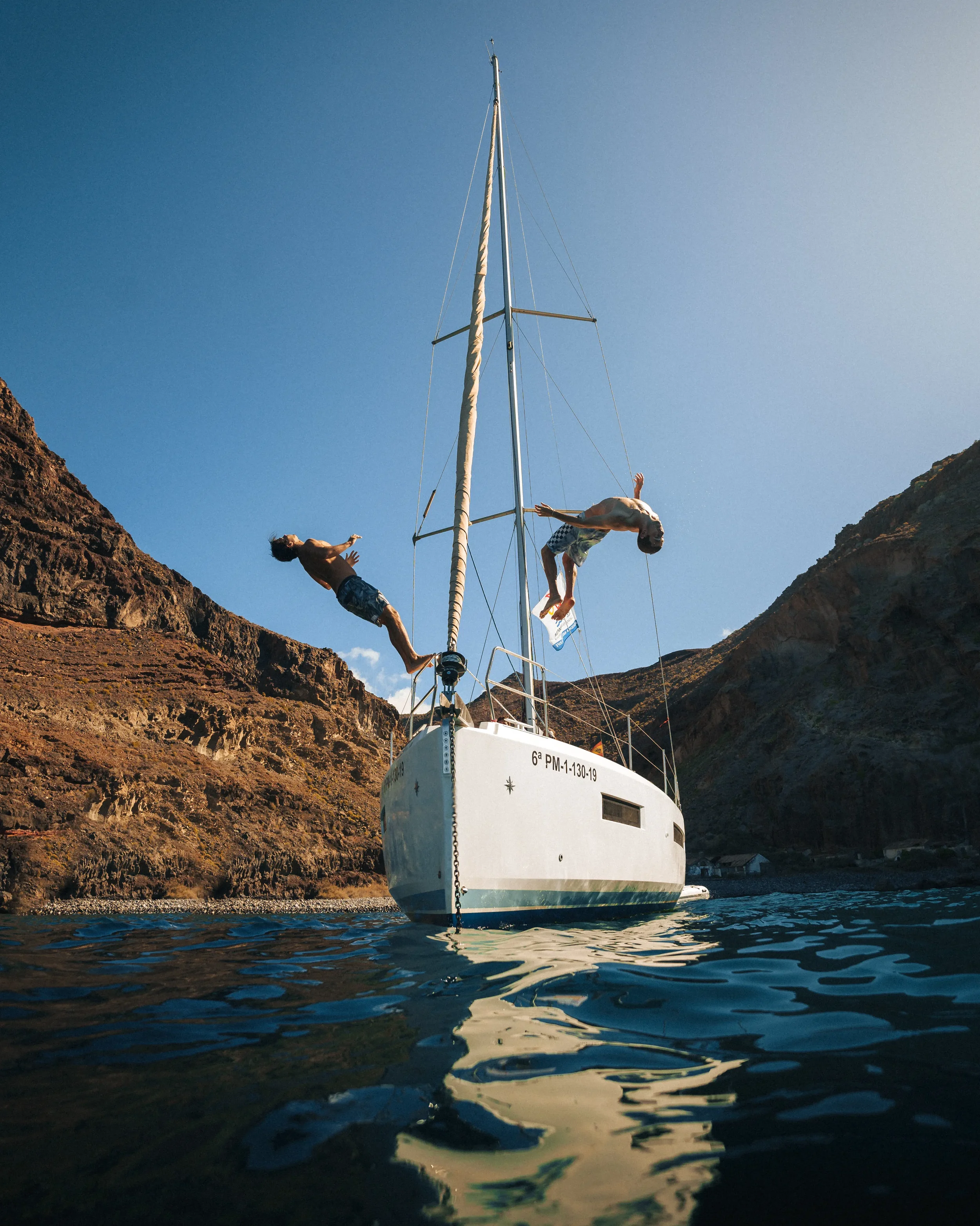 People jumping from a sailboat into the water near rocky cliffs on a sunny day.