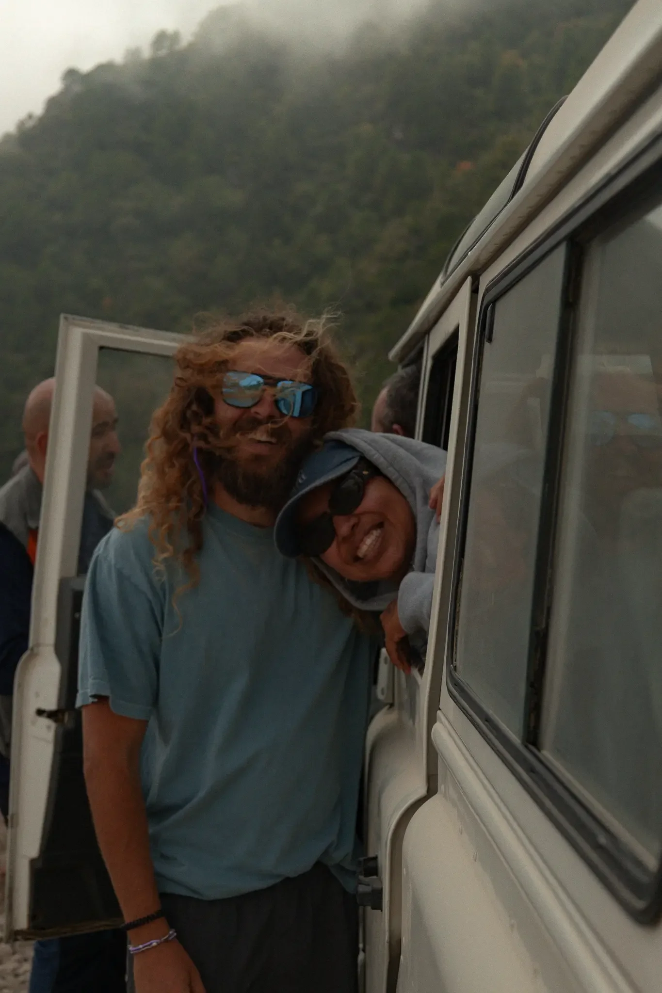People standing outside a cream-colored vehicle with a mountain in the background. One person is smiling and wearing sunglasses, others are visible but partially obscured.