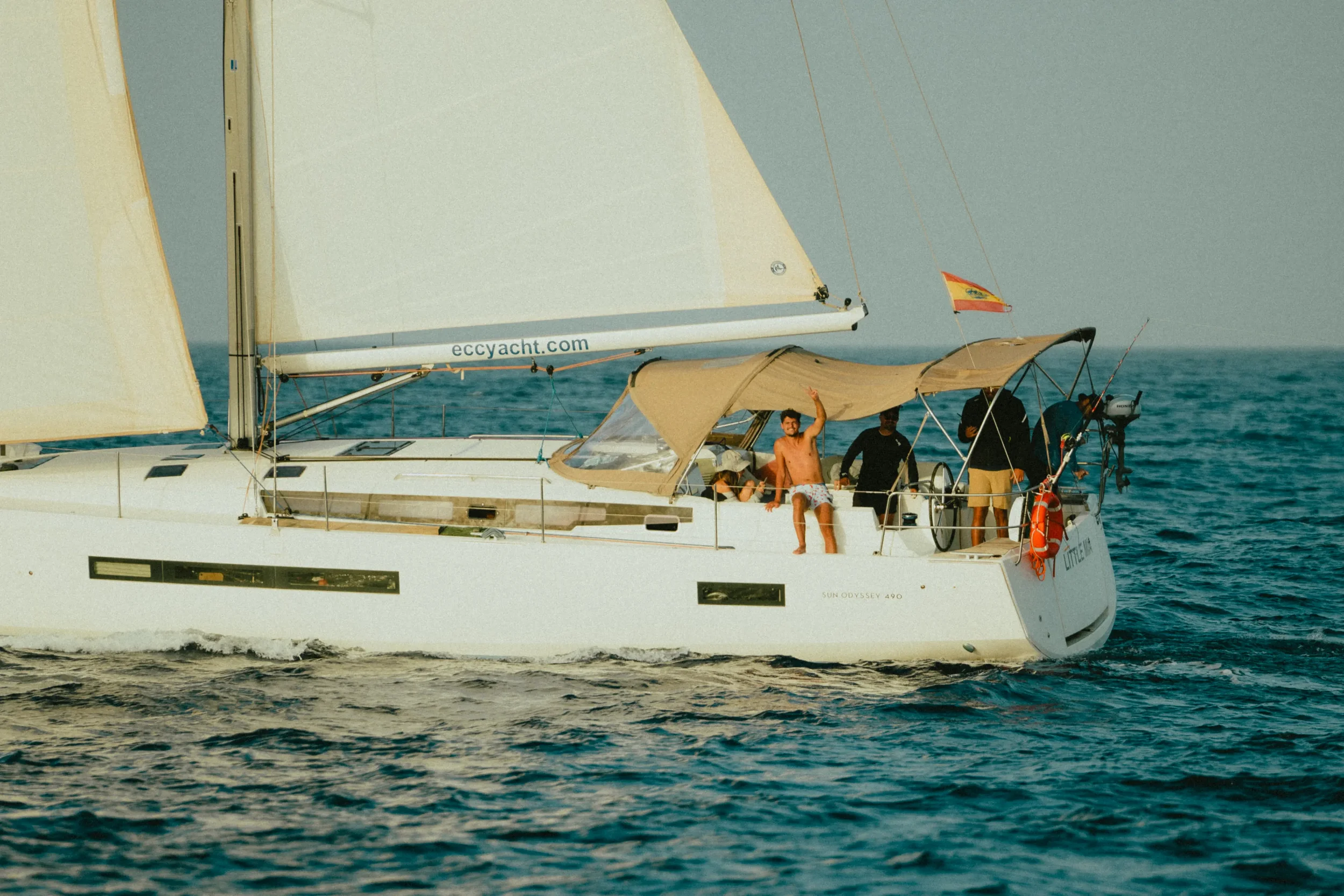 A sailboat on the ocean with people on board, one shirtless man waving, several others sitting or standing, the boat has multiple sails and an orange and yellow flag.