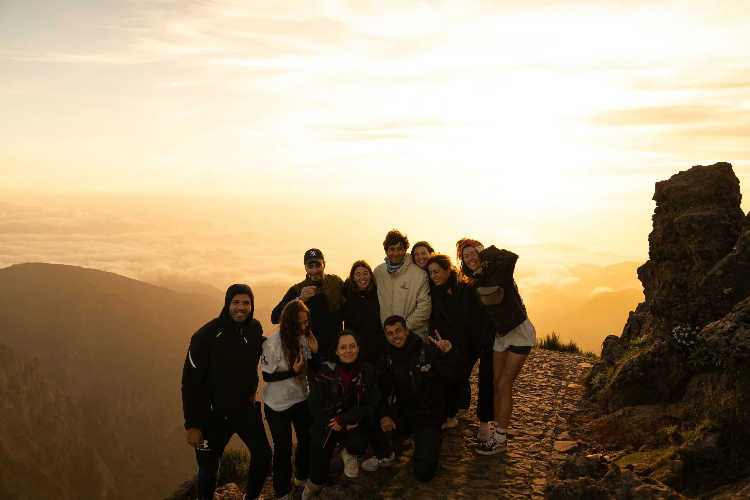 A group of people smiling and posing outdoors during sunset on a mountain trail, with a rocky landscape and cloudy sky in the background.