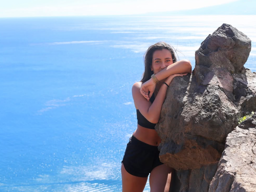A young woman in a black crop top and shorts leaning on a large rock on a cliff overlooking a body of water with a clear blue sky.