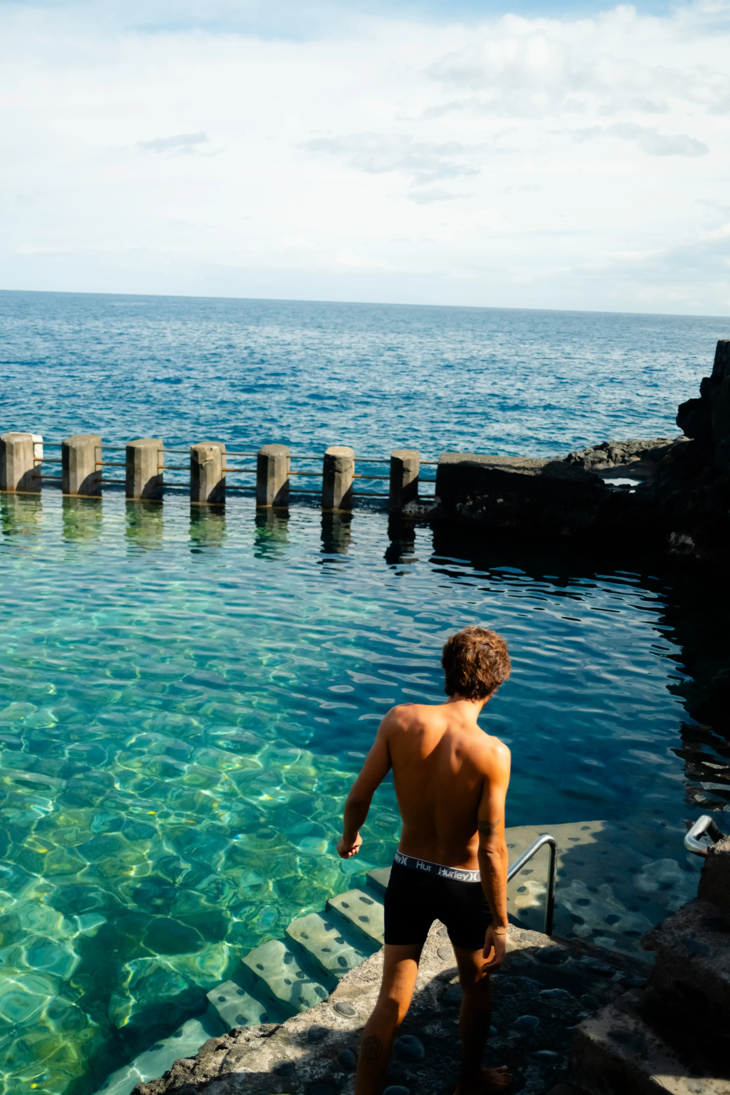 A man with curly hair, wearing black Hurley shorts, stepping into a body of water from a concrete ledge with rocks, overlooking a large ocean with a cloudy sky.