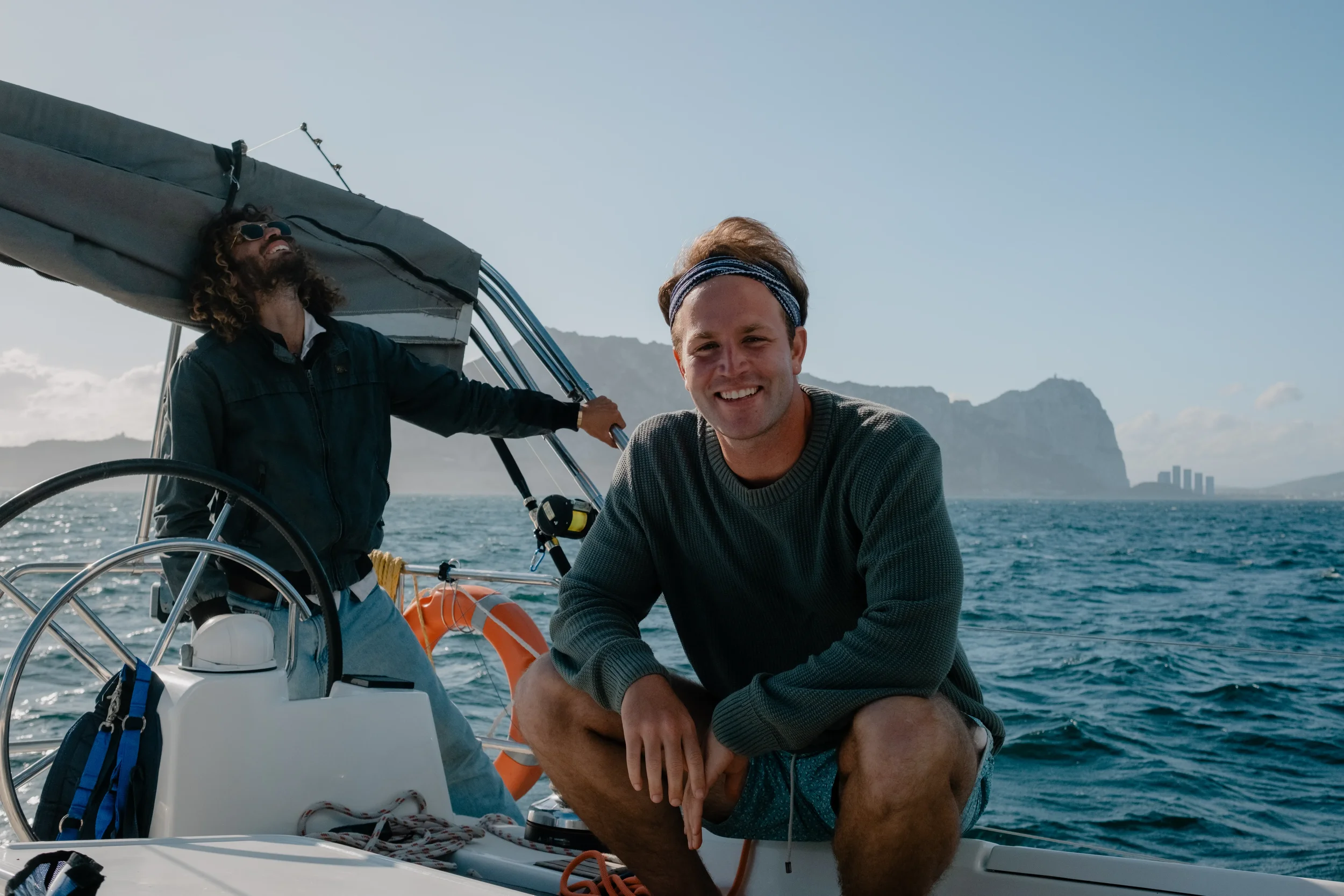 Two men are on a sailboat, one sitting and smiling at the camera, the other standing behind the wheel, looking up and smiling, with mountains and water in the background.