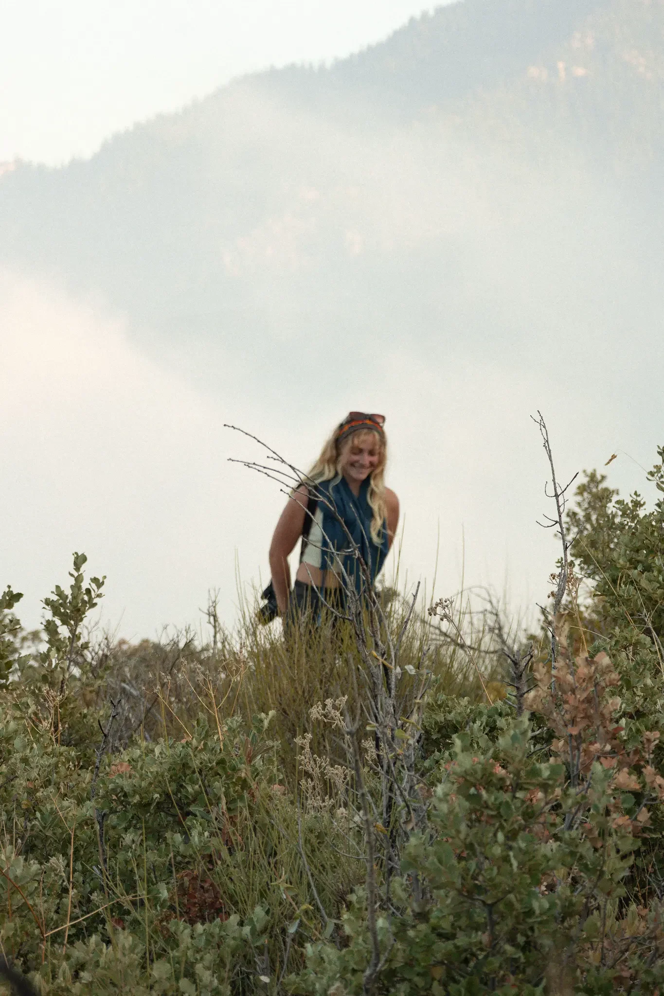 A woman with blonde hair wearing a headband and sleeveless top is standing in a grassy, bushy area with mountains in the background, appearing to be outdoors.