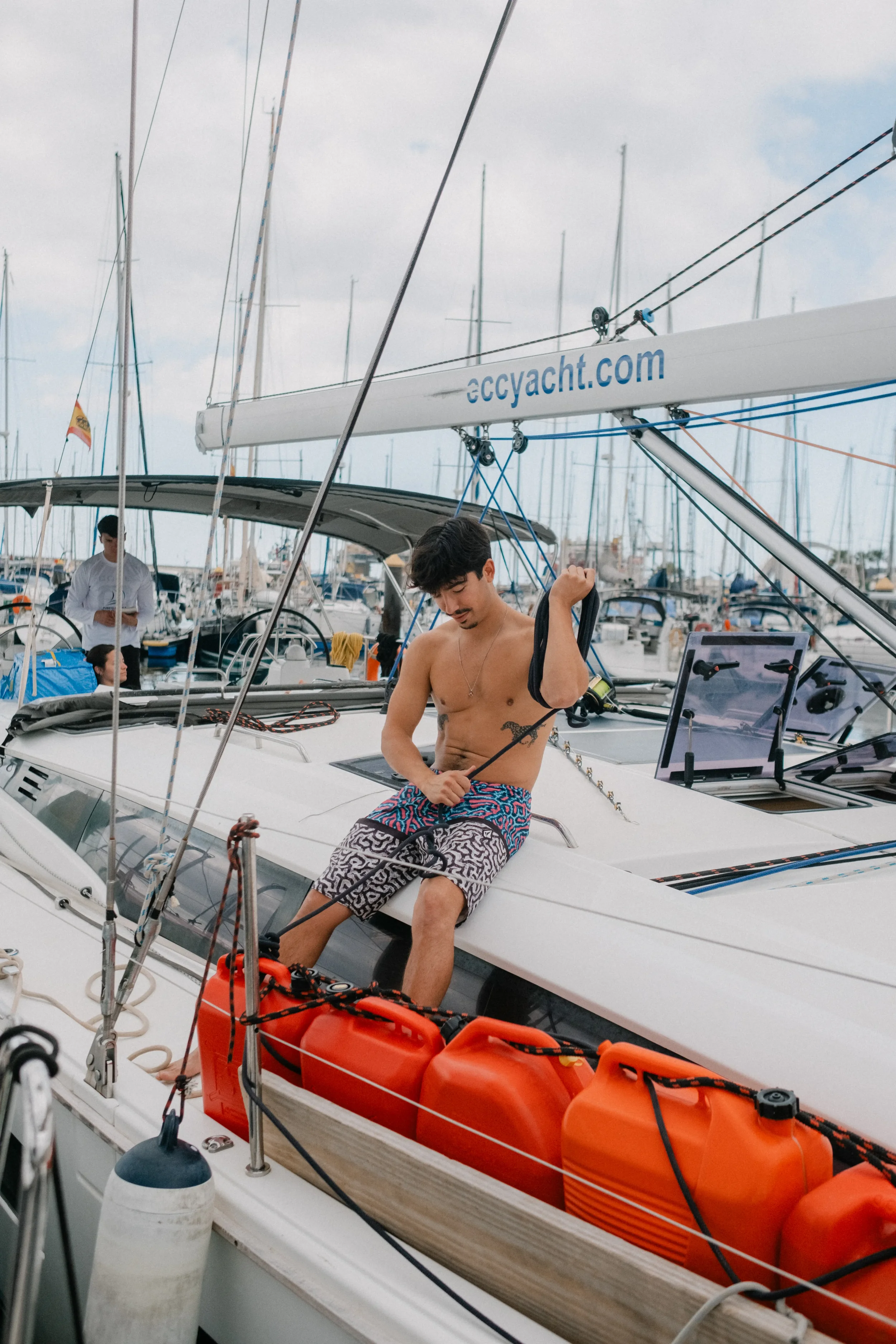 A man with dark hair and a mustache, shirtless and wearing patterned shorts, is sitting on the side of a sailboat in a marina, holding a black strap with a red container nearby, with a young woman sitting in the background.