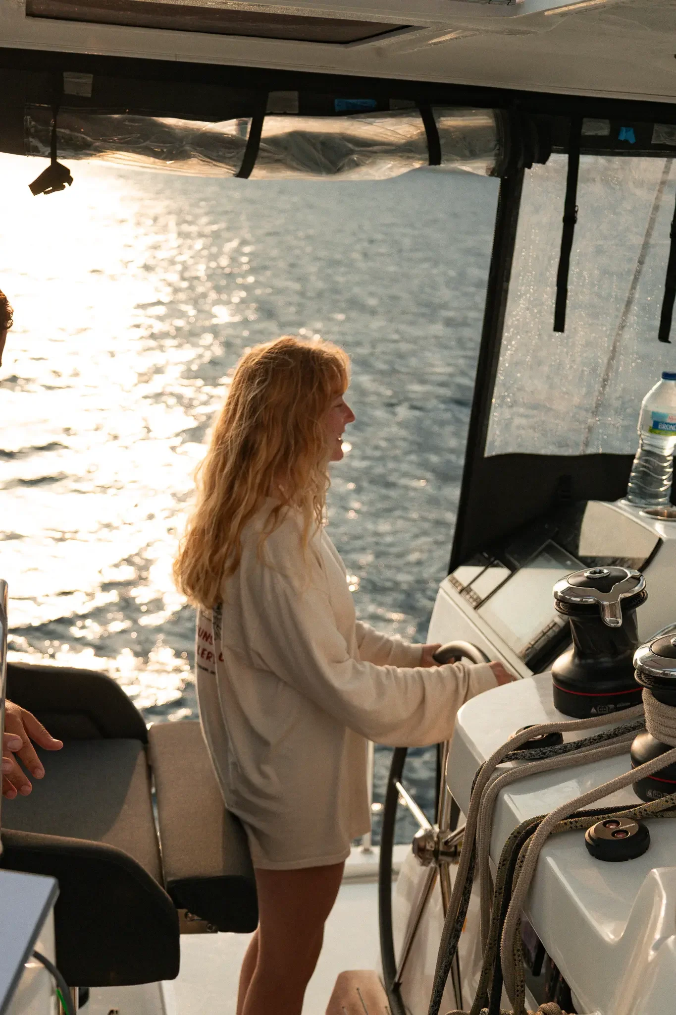 A woman with long blonde hair on a boat, smiling and wearing a light-colored hoodie and shorts, with water and the setting sun in the background.