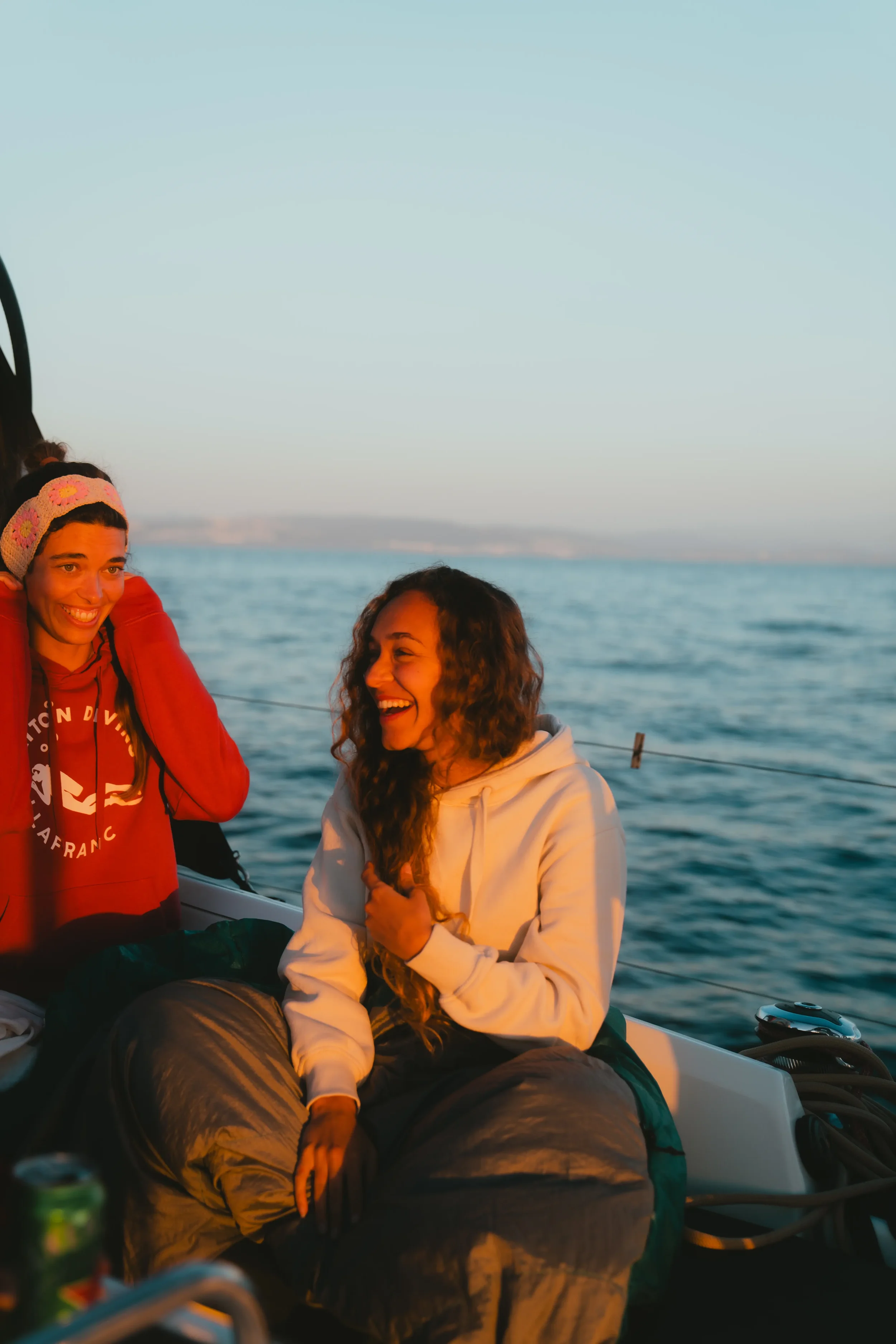 Two women sitting on a boat, one with curly hair in a white hoodie and the other with straight hair in a red hoodie with a headband, enjoying each other's company with water in the background during sunset or sunrise.