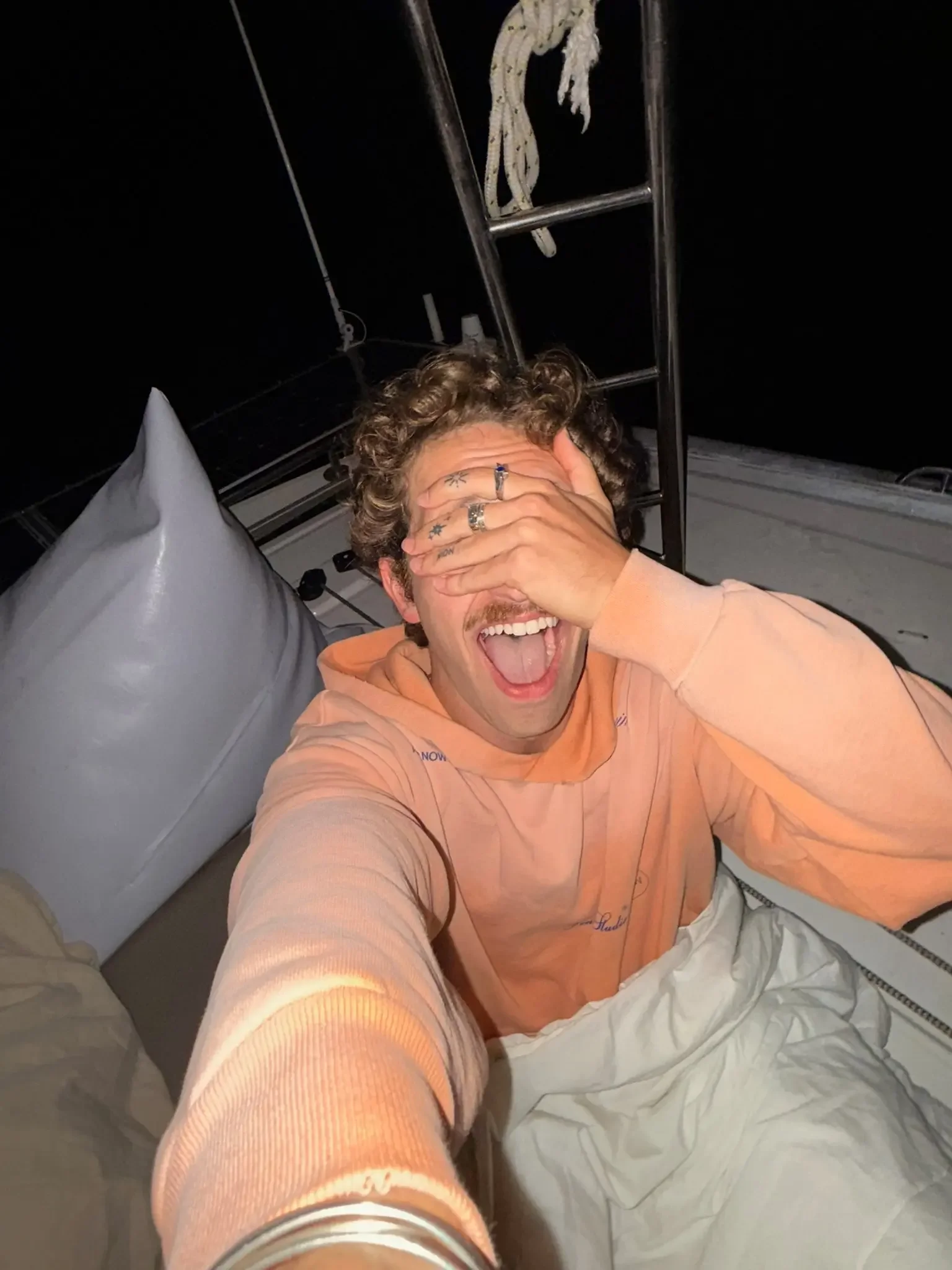 A man with brown, curly hair and facial hair sitting on a bed, wearing a peach-colored hoodie, smiling widely, with his hand covering part of his face. The bed has white sheets and pillows, and there is a ladder and black netting or fabric above him.