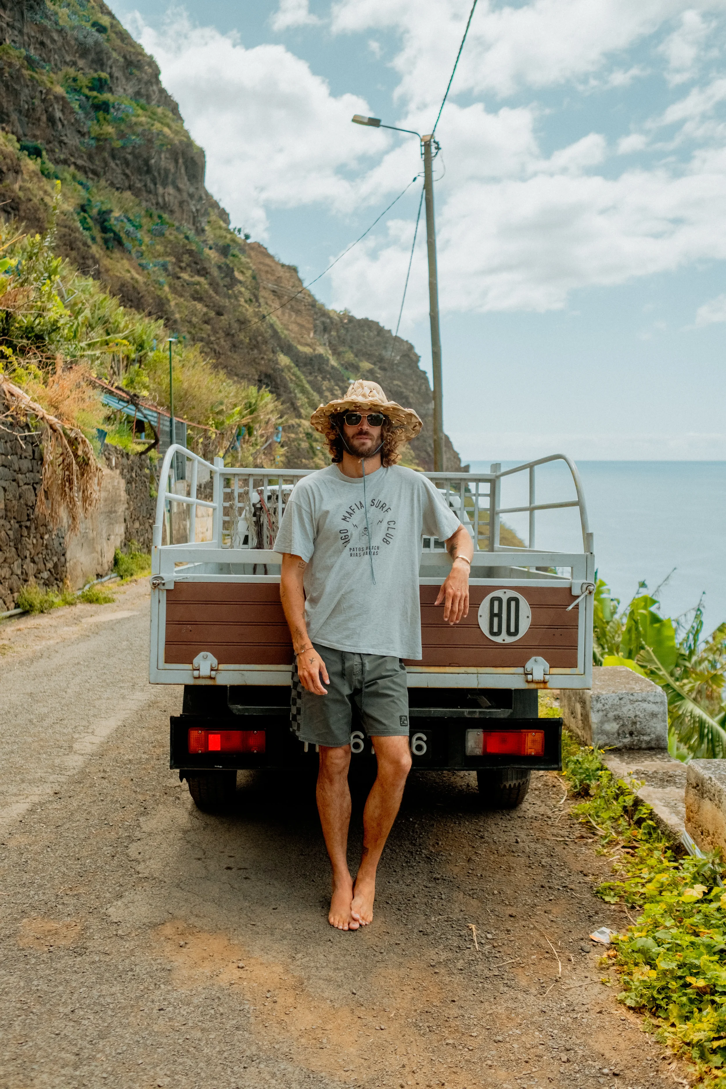 A man with curly hair, wearing sunglasses, a wide-brimmed straw hat, a t-shirt, and shorts, is barefoot and standing in front of a small truck with an open back on a dirt road. Behind him, there is a cliffside with green vegetation and a cloudy sky o