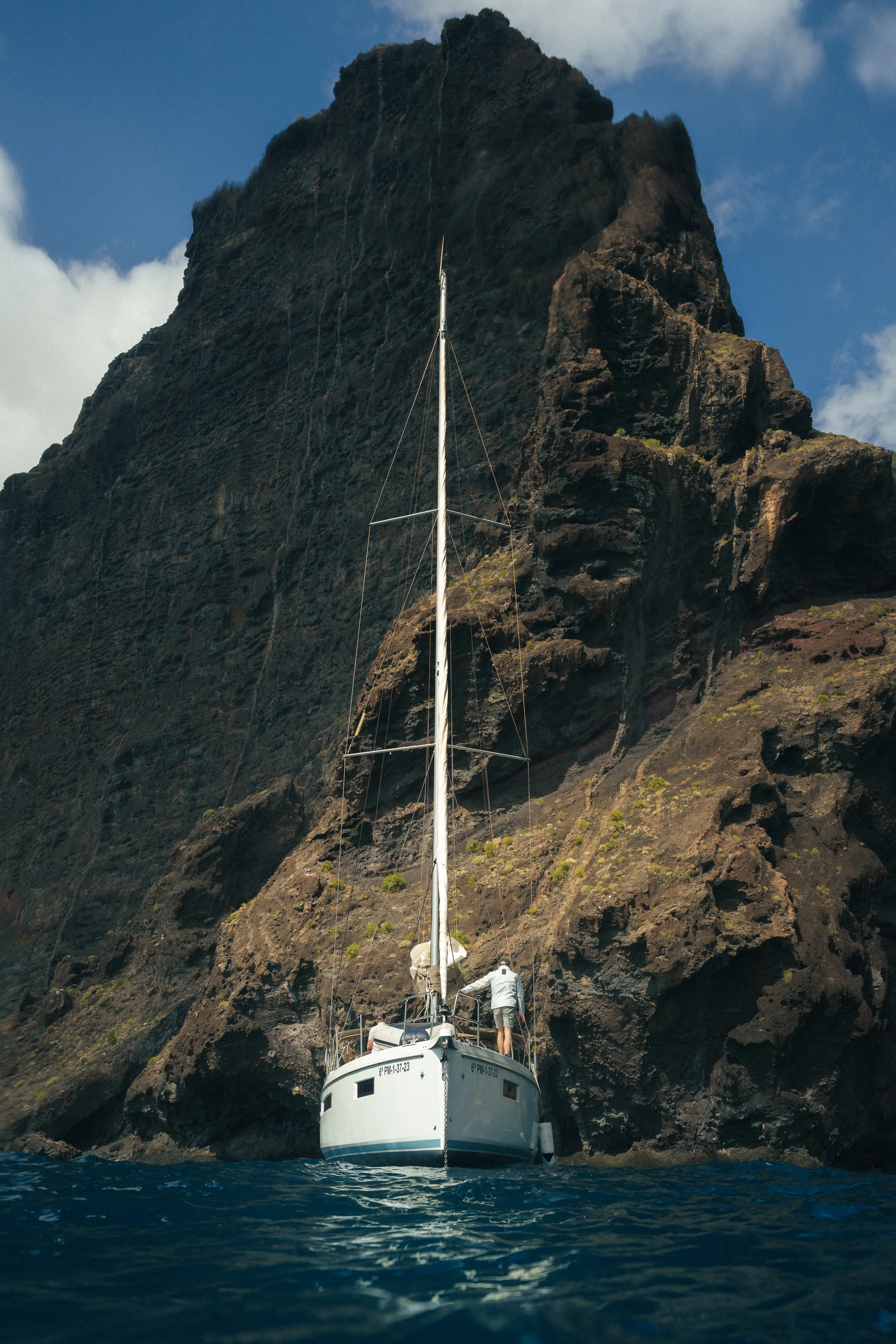 A sailboat is anchored near a towering rocky cliff with a person standing on the deck near the stern, overlooking the water. The sky is partly cloudy.