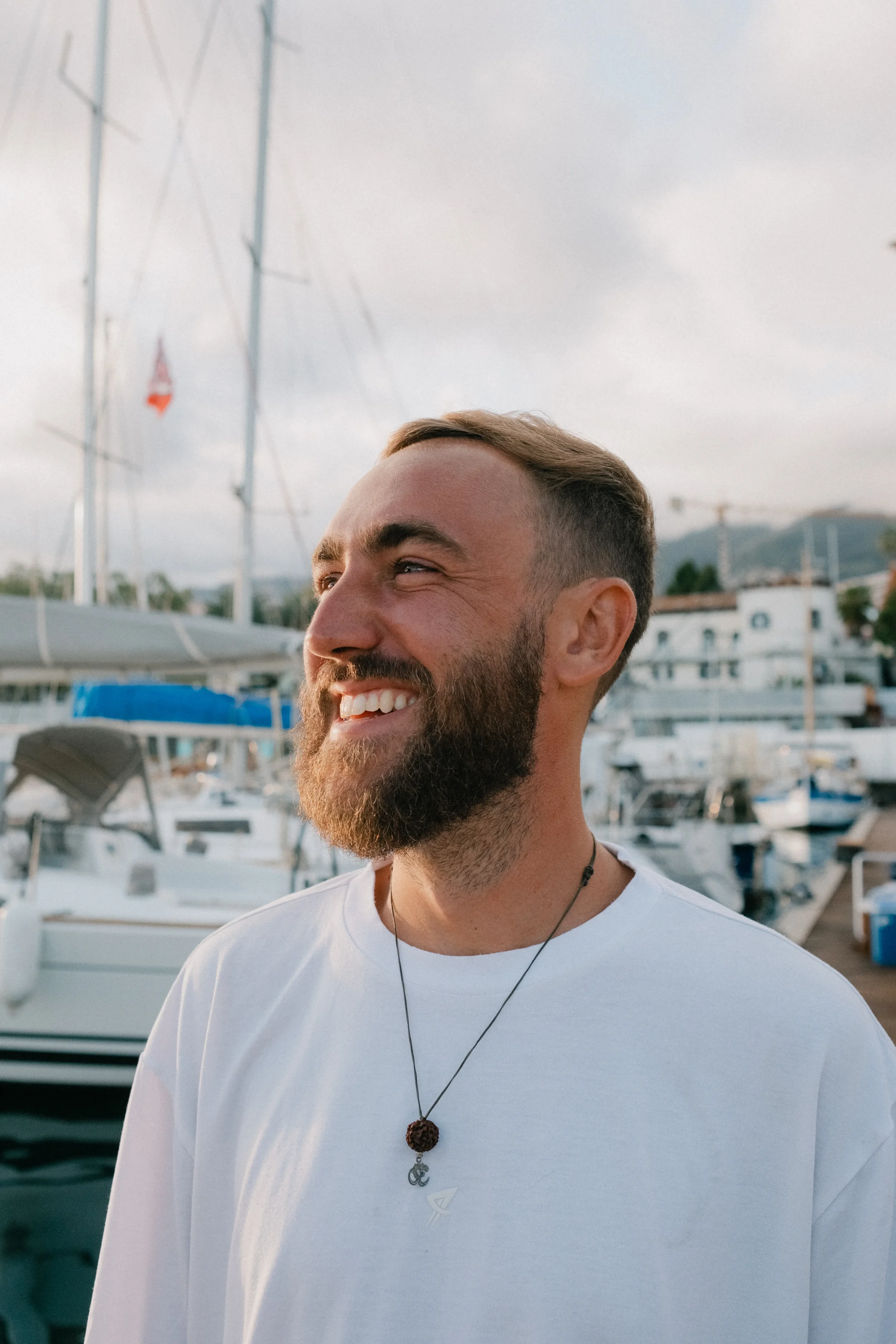 Close-up of a smiling man with a beard and short hair, wearing a white T-shirt, a necklace with a seashell and other charms, standing near a marina with boats and a cloudy sky in the background.