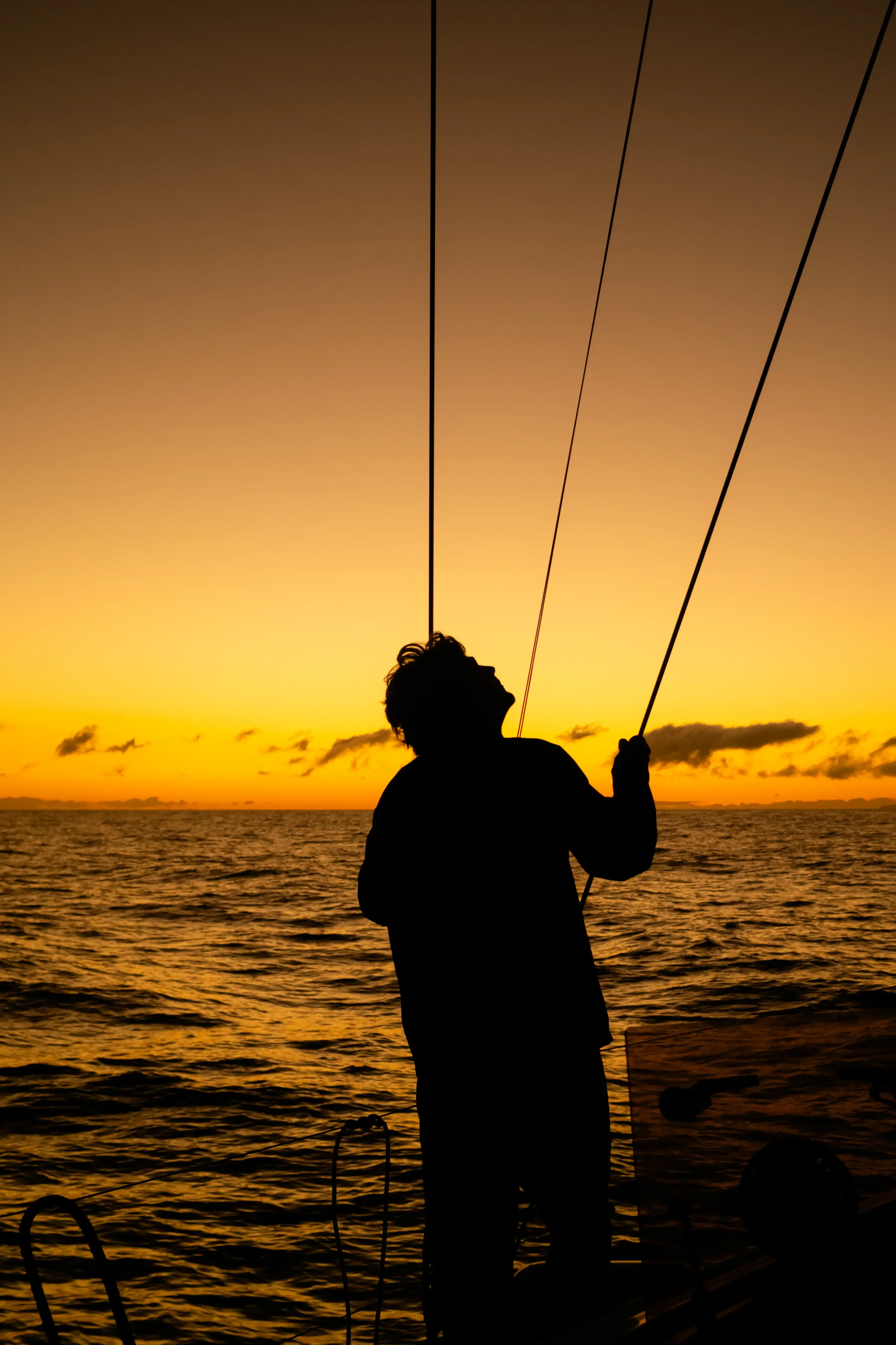 A person with curly hair stands on a boat, holding onto ropes, silhouetted against a vibrant orange and yellow sunset over the ocean, with a few clouds in the sky.