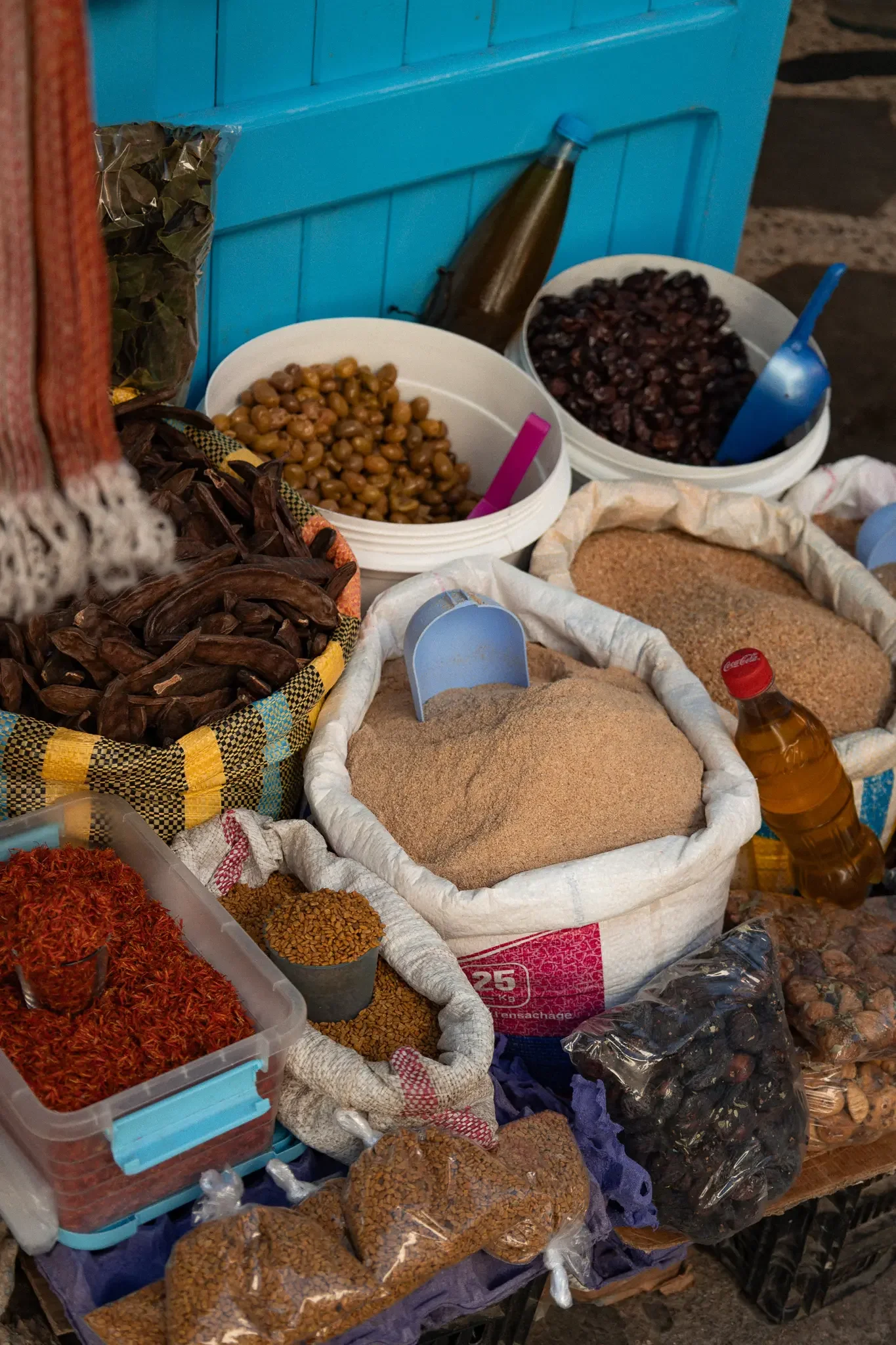 A market stall displaying various dried goods such as beans, spices, and seeds in plastic containers and bags, with a blue wooden wall in the background.