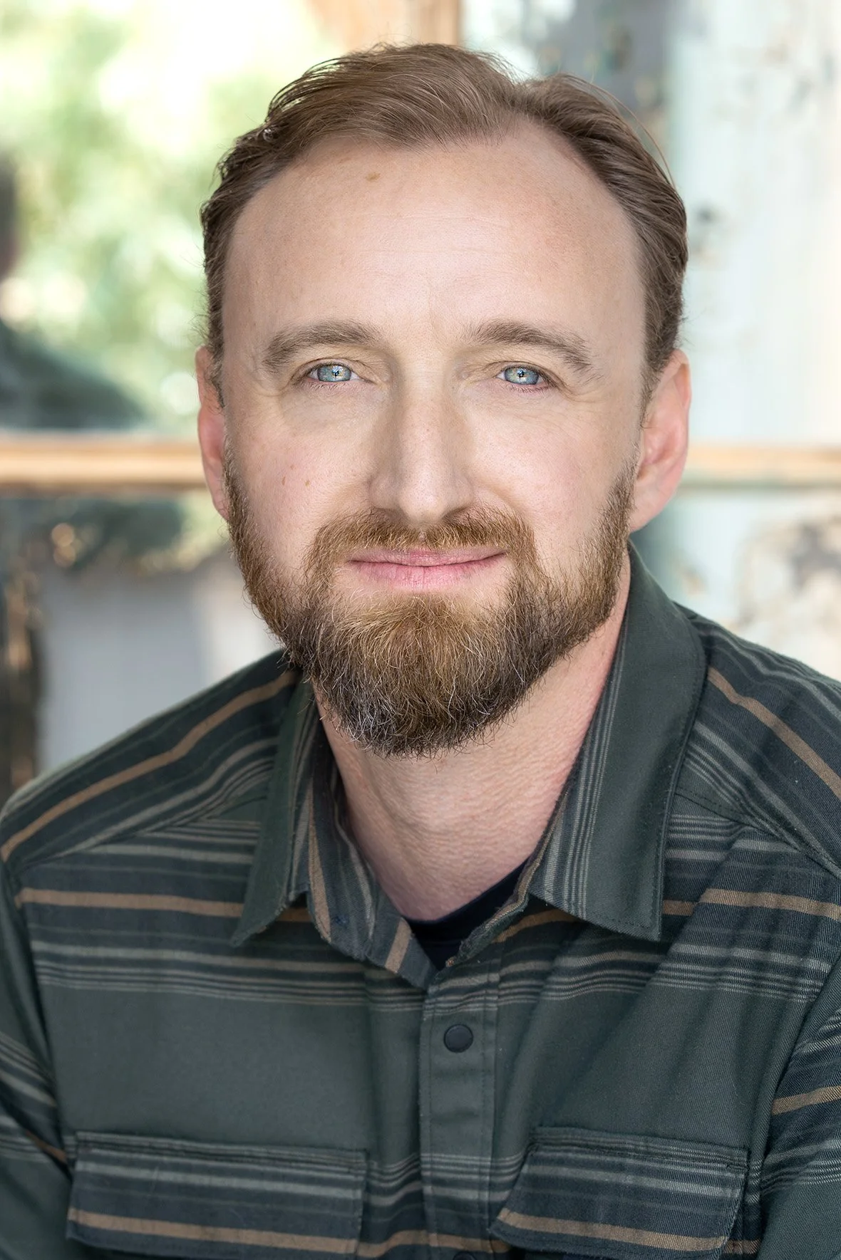 A man with blue eyes and a beard, smiling, in a striped shirt, sitting indoors with blurred background of windows and greenery.