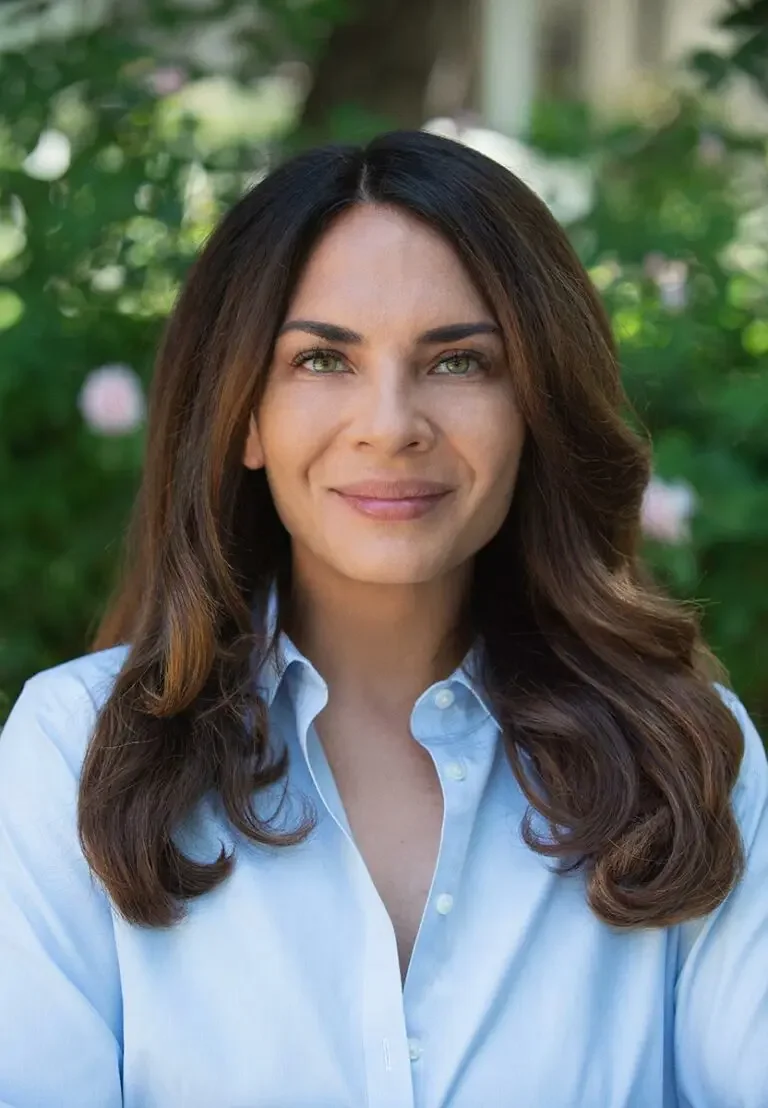A woman with brown hair and green eyes smiling outdoors, wearing a light blue button-up shirt.