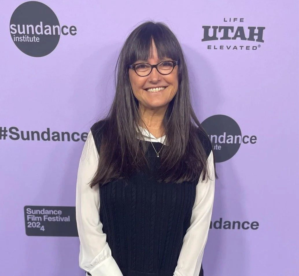 Woman with long brown hair and glasses smiling at the camera, standing in front of a purple backdrop with Sundance Institute, Sundance Film Festival 2024, and Utah Elevated logos.