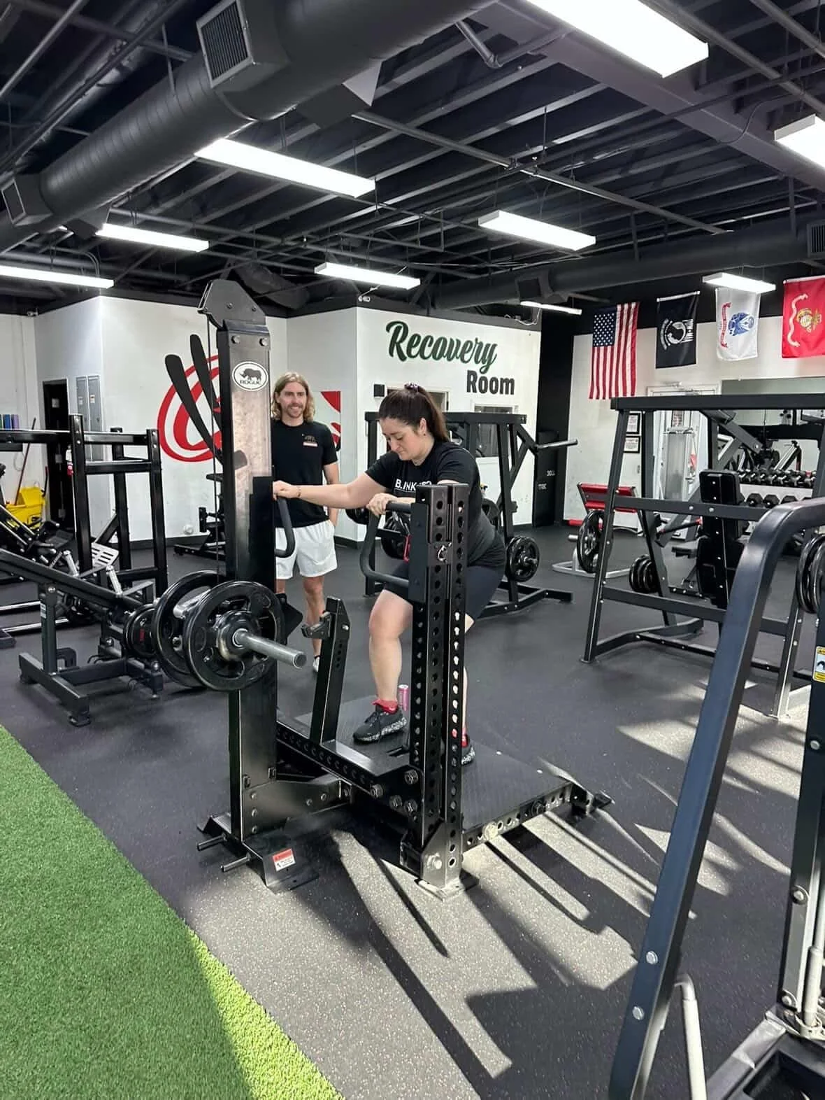 A woman lifting weights on a machine in a gym, with a man standing nearby and a sign that says 'Recovery Room' in the background.