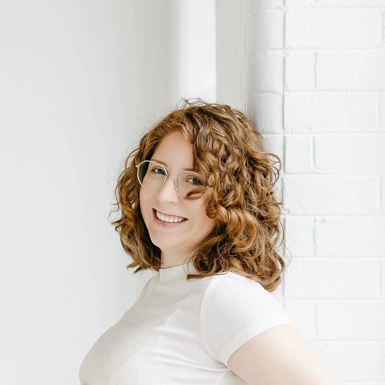 A woman with curly red hair and glasses smiling while leaning against a white brick wall.