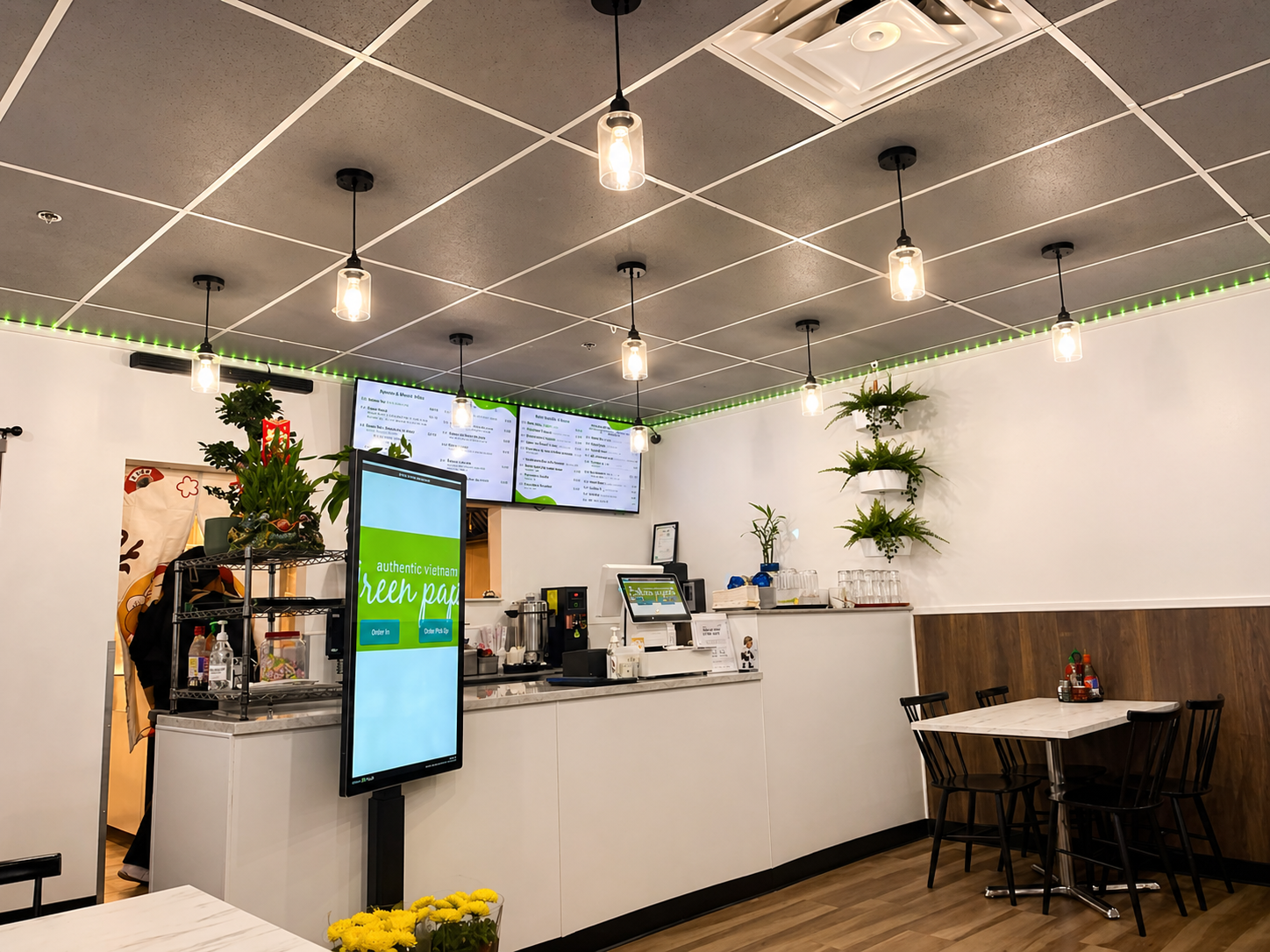 Inside a modern Vietnamese restaurant with a counter area, digital menu screens, and seating for customers. Decor includes green plants on the walls, hanging lights, and a small table with condiments.