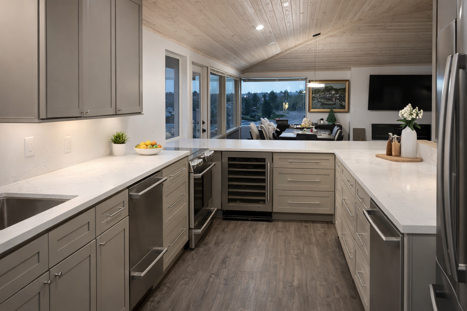 Modern kitchen with grey cabinets, white marble countertops, stainless steel appliances, and a view into the dining and living area with large windows and a scenery outside.