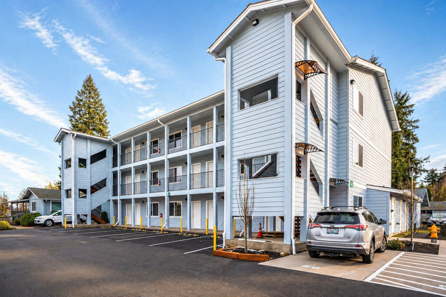 Exterior view of a light blue multi-story apartment building with balconies, black railings, and a parking lot with several cars, including a gray Toyota SUV, under a sky with scattered clouds.